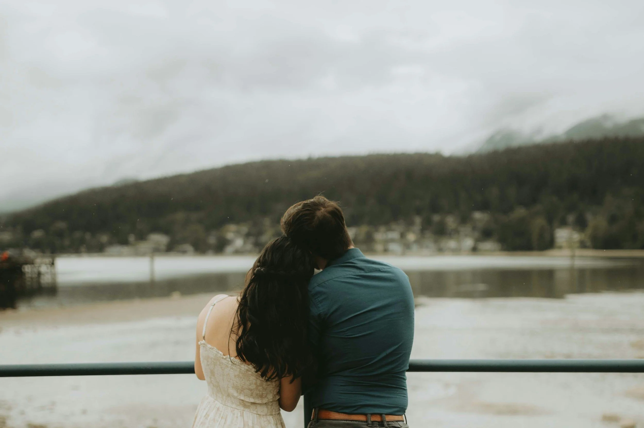 A couple sits close together on a railing, facing a lake with mountains and supporting each other.