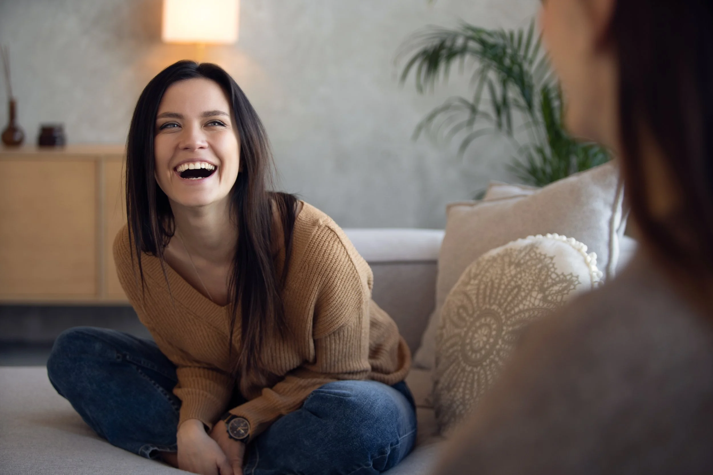 A woman sitting outdoors by the water during sunset, facing away from the camera, with her hair blowing in the wind.