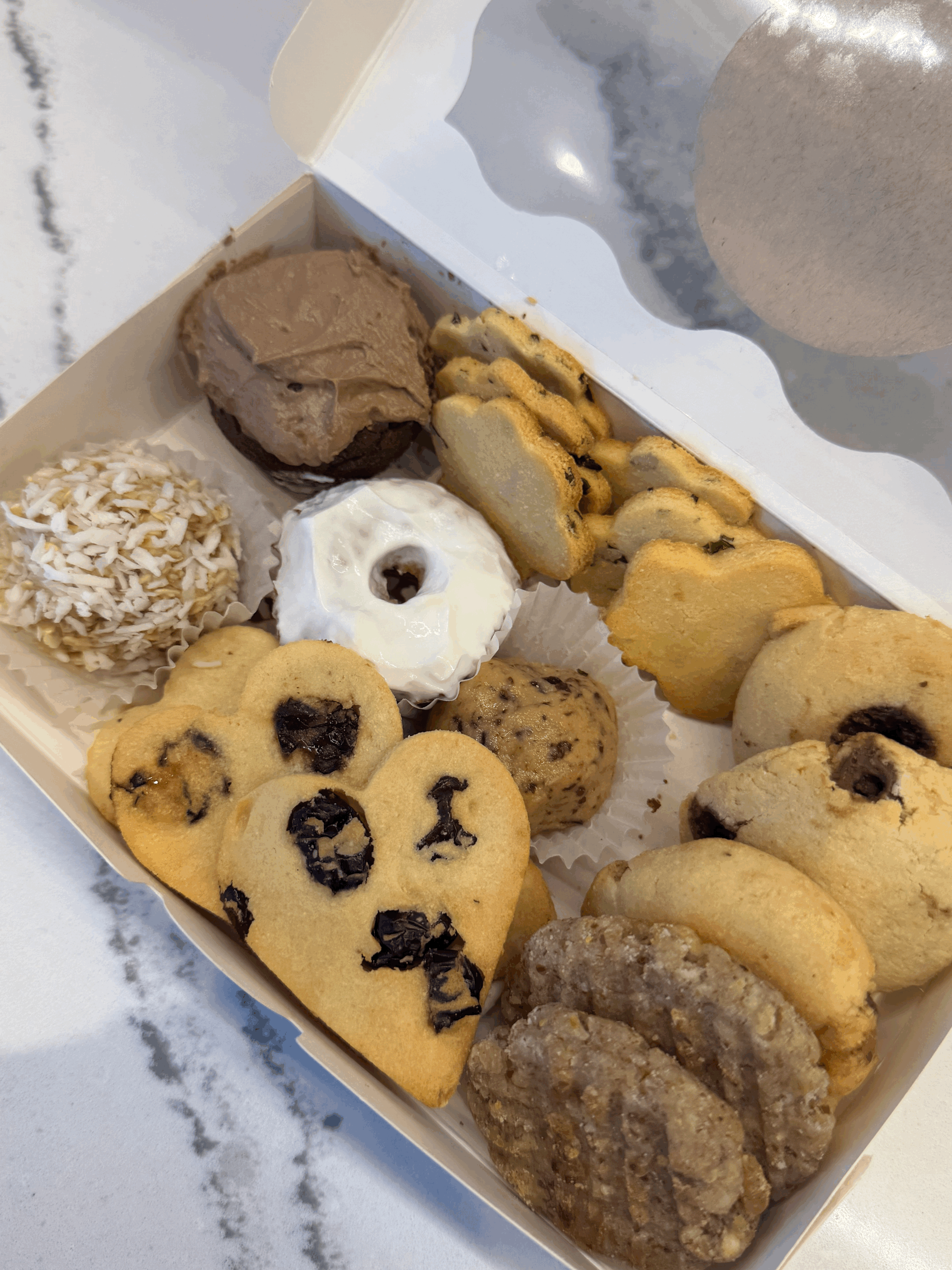 Assorted dog treat box with a variety of fun-shaped, all-natural treats including mini cupcakes, carob cookies, doughnuts, and heart-shaped biscuits