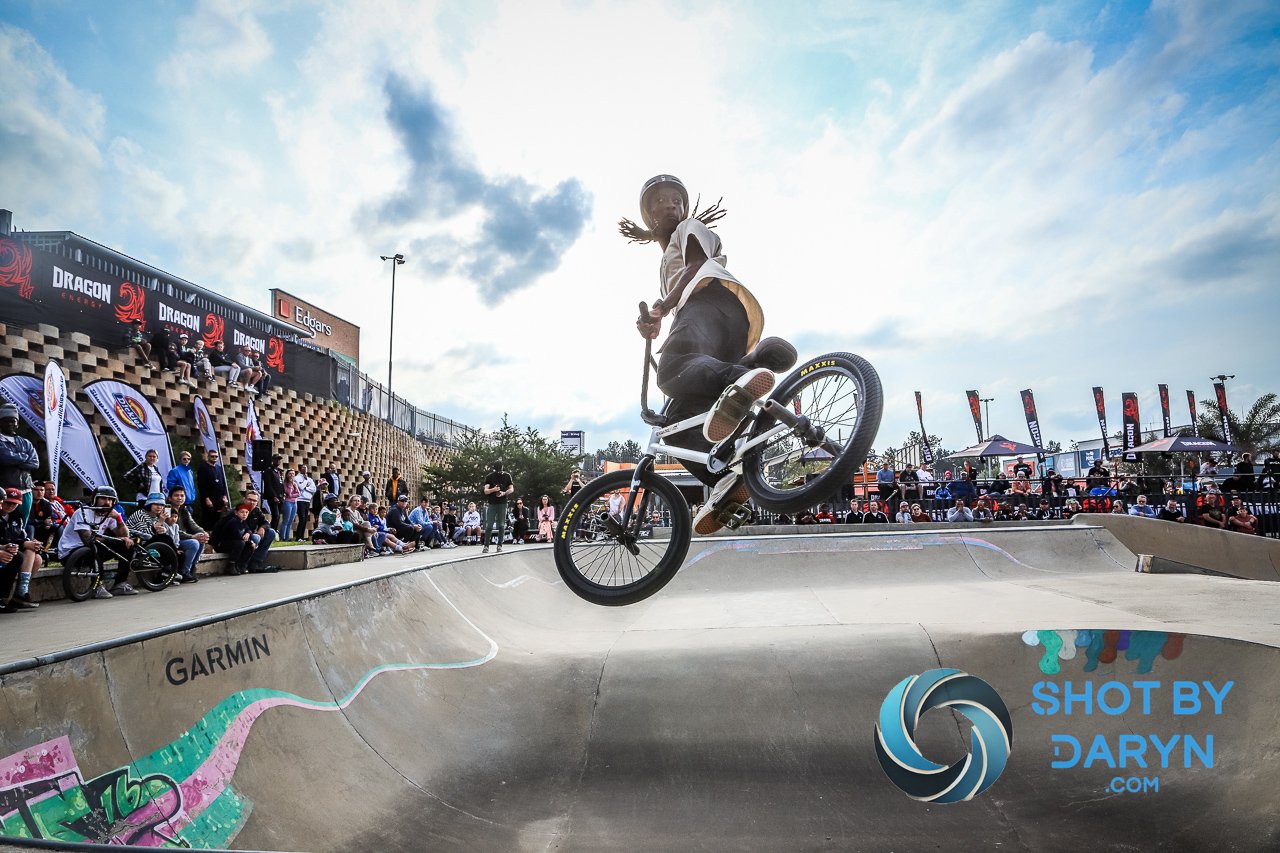 BMX rider performing a trick in a skate park bowl with spectators watching. The sky is partly cloudy, and there are banners and advertisements in the background. The event appears to be well-attended.