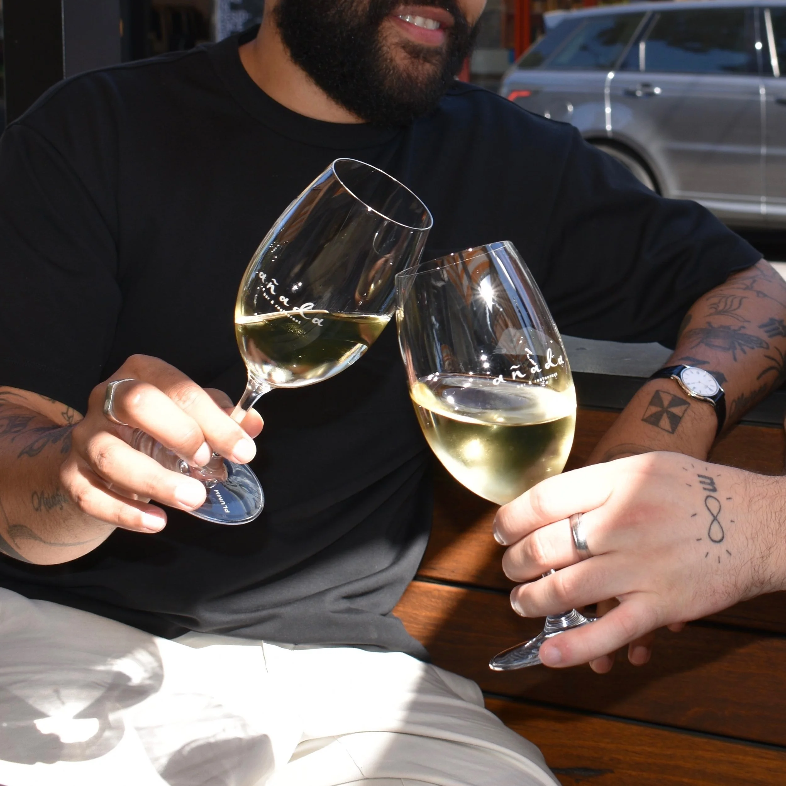 A man with tattoos on his arms holding two glasses of white wine, clinking them together while sitting at a wooden table.