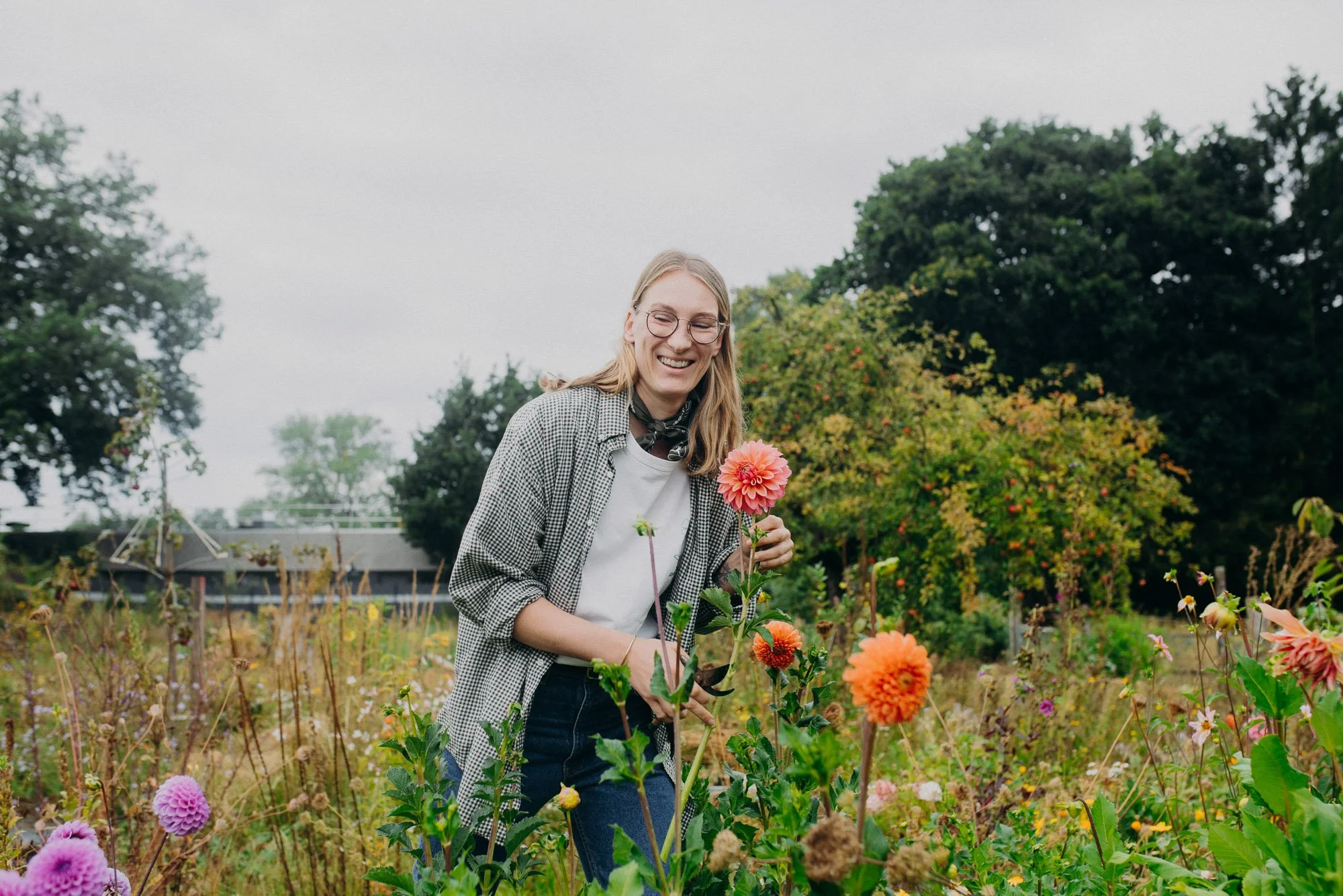 A woman with glasses smiling and holding a pink dahlia flower in a garden filled with various colorful flowers, with trees and a building in the background.