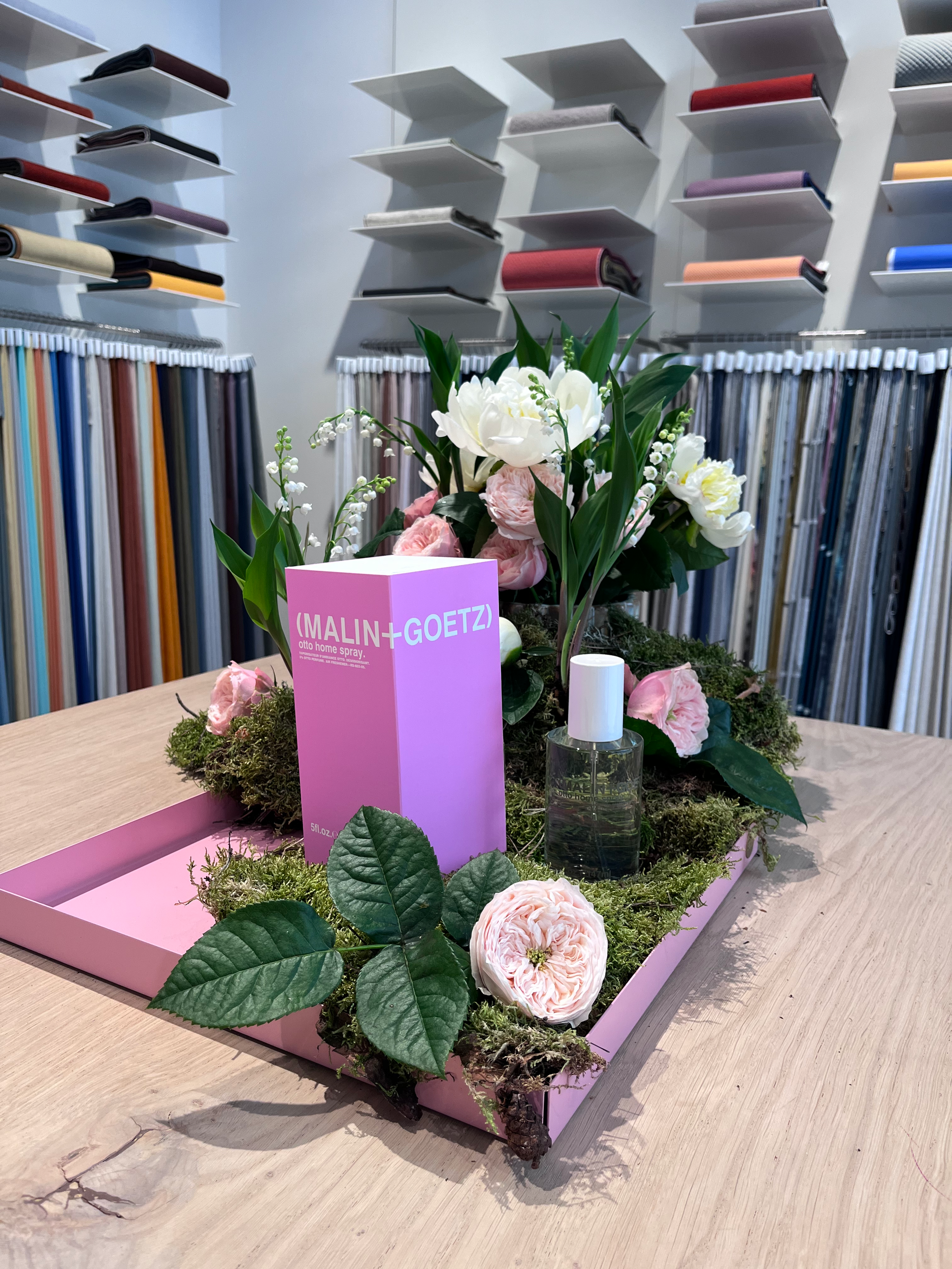 A decorative display featuring a pink Malin + Goetz perfume box, a small bottle of fragrance, and fresh pink and white flowers with green leaves, arranged on a pink tray with moss, on a wooden table in a fabric store or upholstery shop.