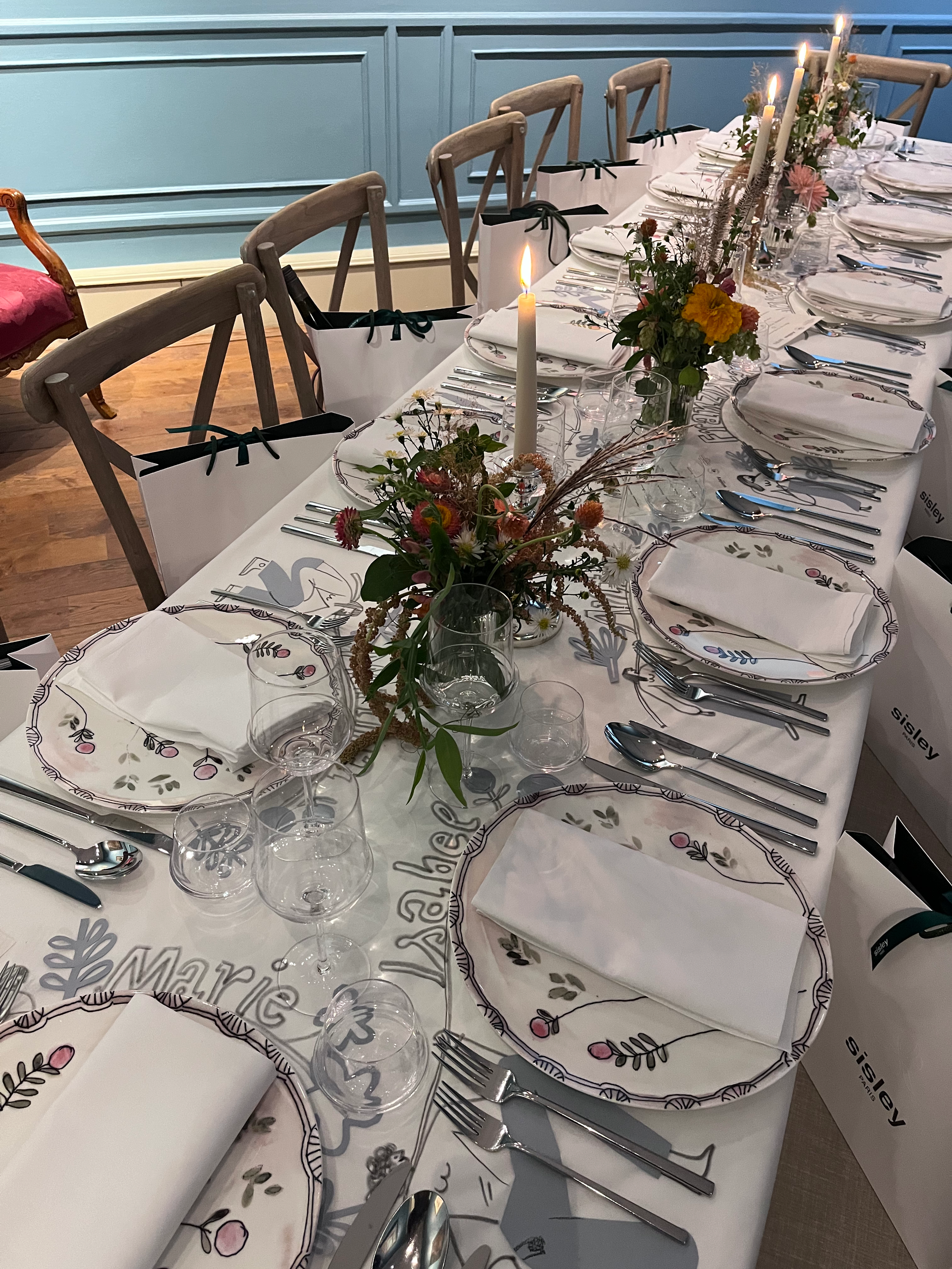 A long dining table set with white plates, napkins, and silverware, decorated with floral centerpieces and tall candles, with gift bags placed along the chairs, in a room with blue walls and wooden flooring.