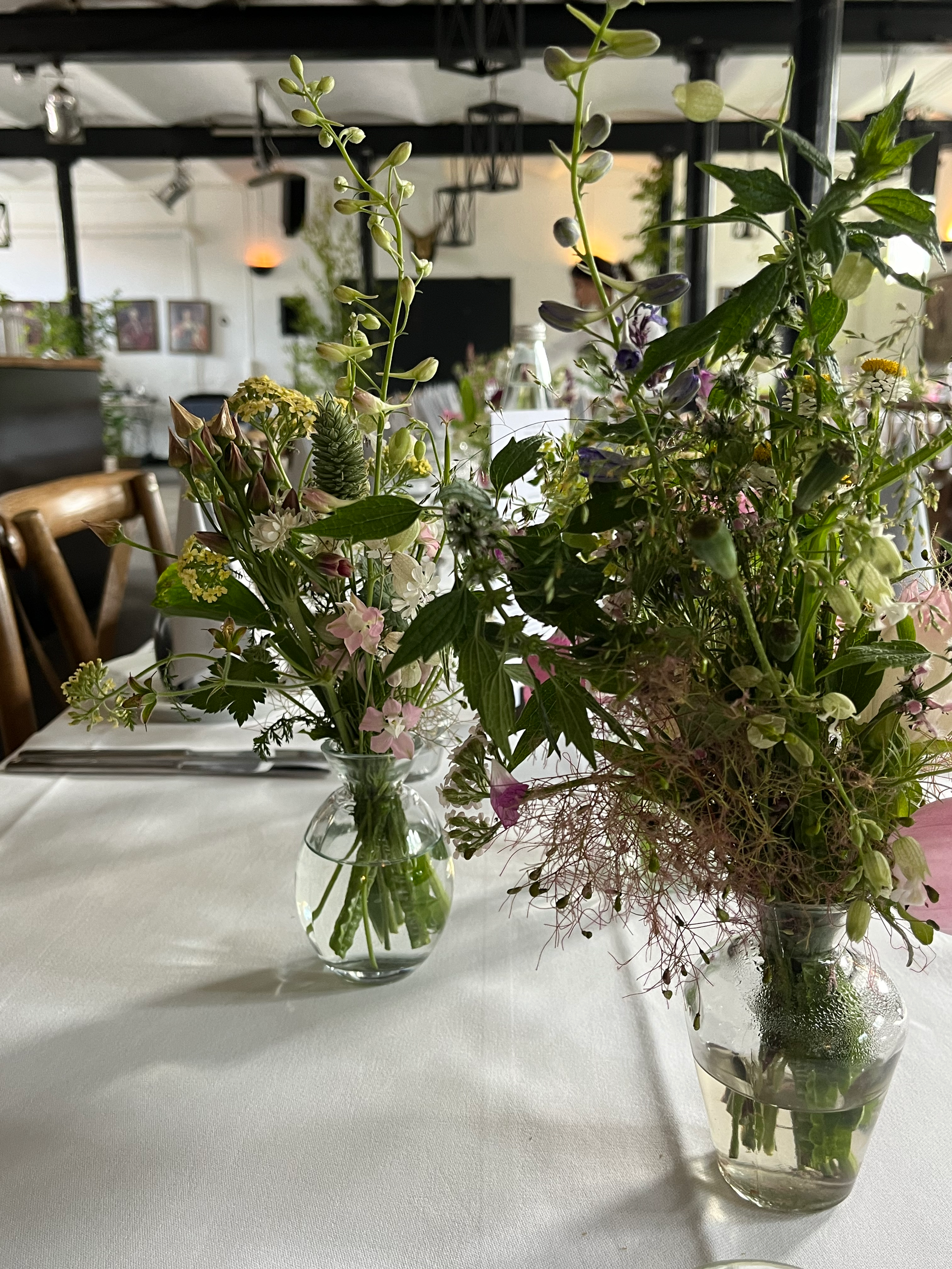 Two glass vases with fresh flowers on a white tablecloth in a restaurant or event space, with a blurred background of chairs and interior decor.