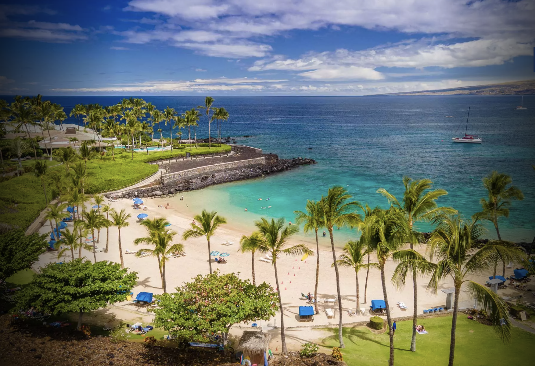 A tropical beach with white sand, blue-green water, and palm trees. There are umbrellas, chairs, and people relaxing on the beach, with a lush green landscape and a stone wall in the background. The ocean and sky are visible in the distance.