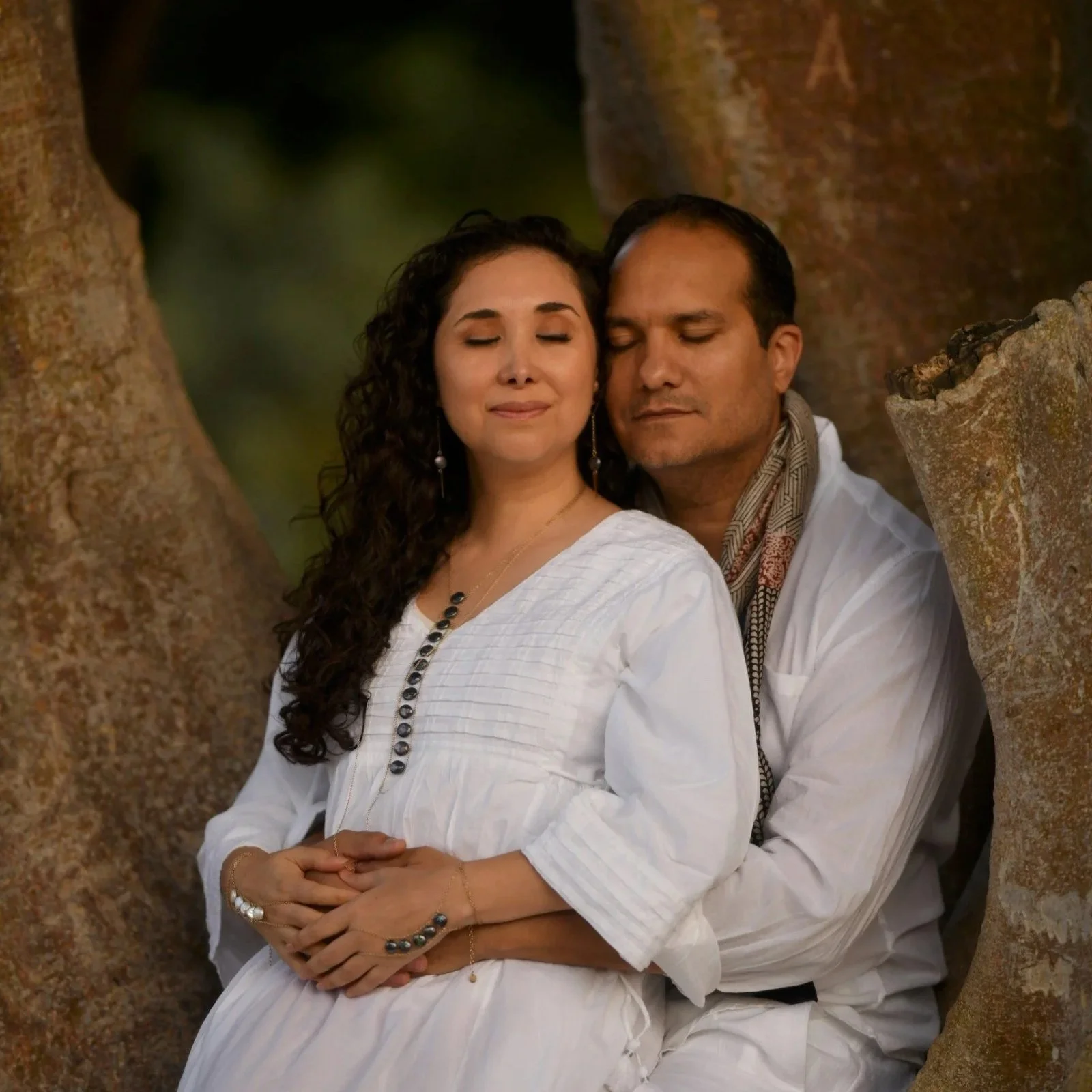 Woman in tan dress kneeling on a carpeted floor, hands forming a heart shape over her lap, with curly hair and decorative necklace.