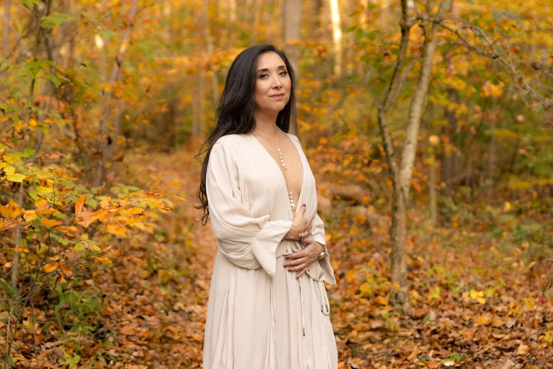 Woman in a cream-colored dress in a forest with autumn leaves.