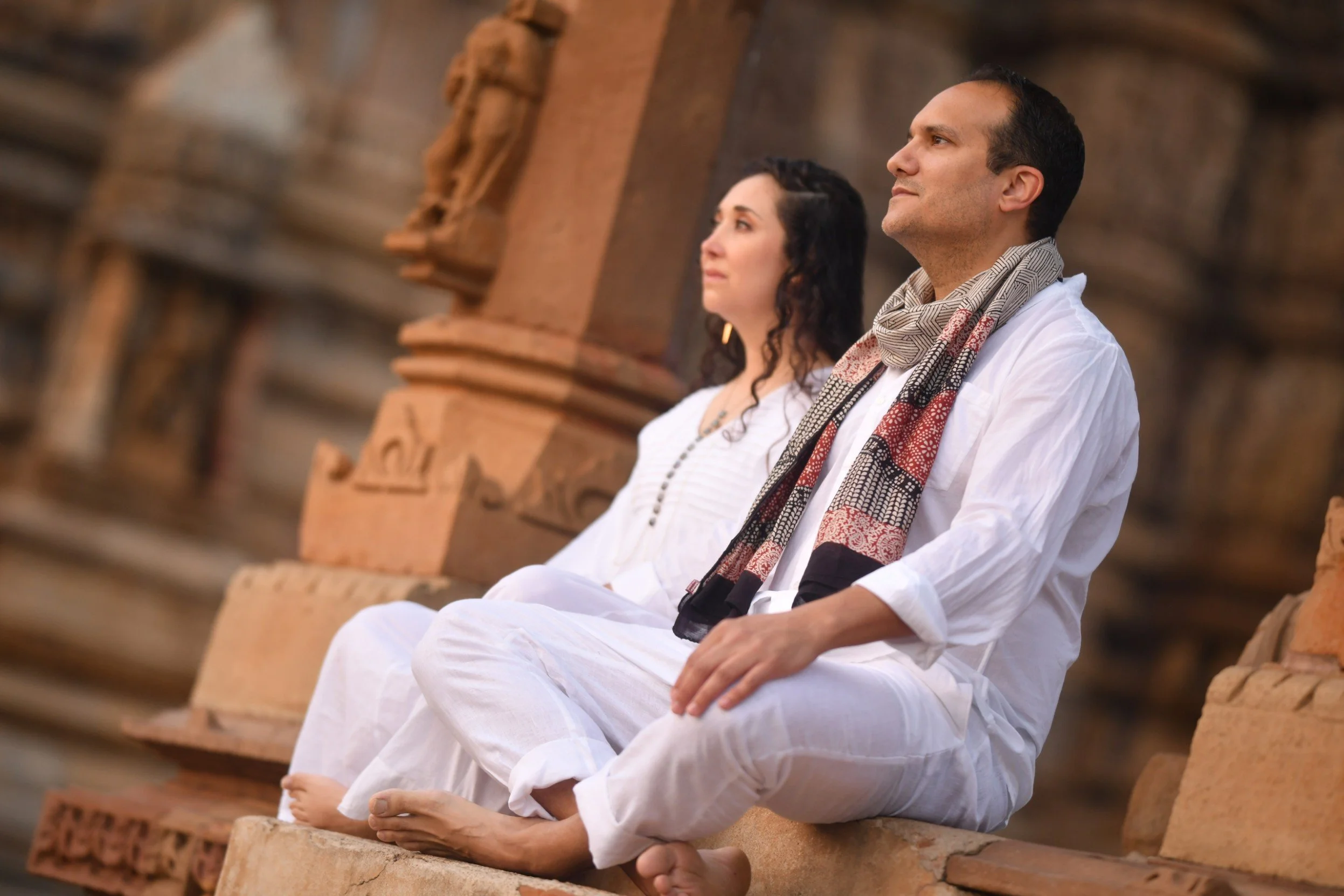 Woman in tan dress kneeling on a carpeted floor, hands forming a heart shape over her lap, with curly hair and decorative necklace.
