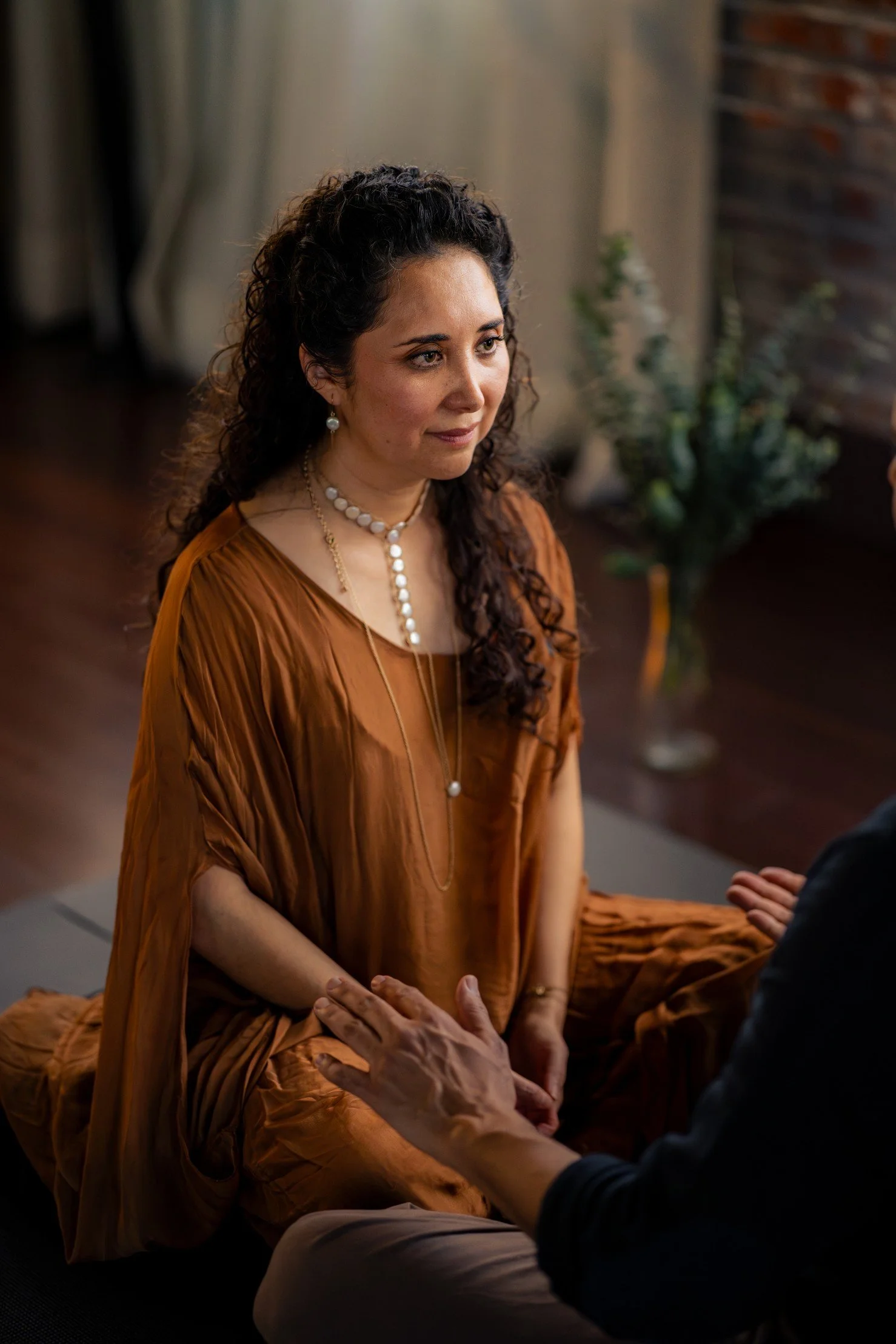Woman in tan dress kneeling on a carpeted floor, hands forming a heart shape over her lap, with curly hair and decorative necklace.