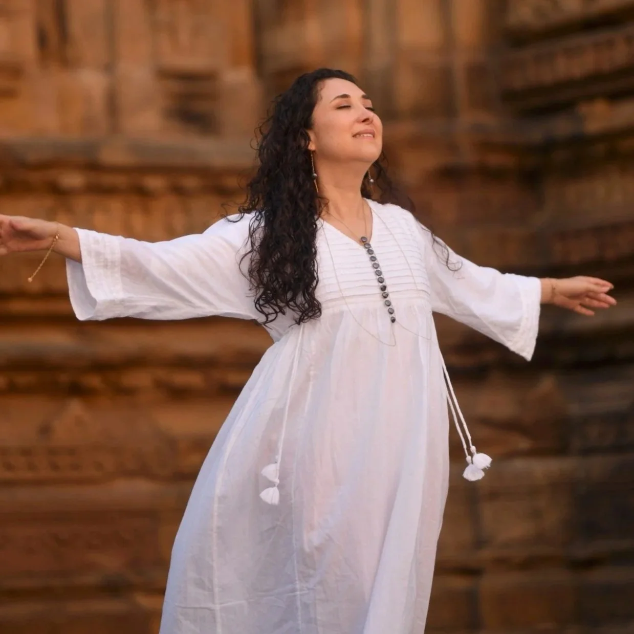 A woman with curly hair is sitting on a couch, writing in a notebook. She is wearing a beige dress and jewelry, and is surrounded by decorative pillows and indoor plants. A window is visible in the background.