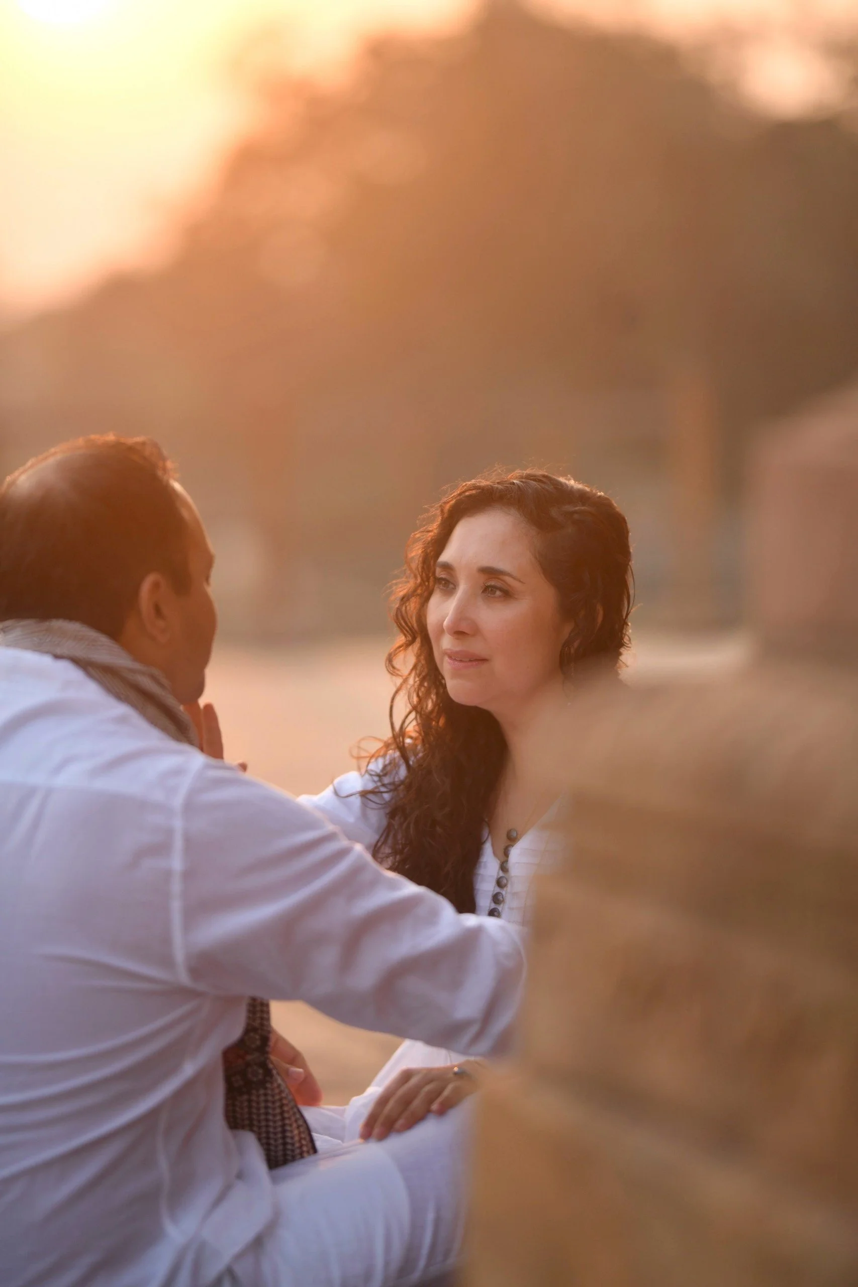Woman with curly hair and eyes closed, wearing a ruffled top, posed indoors with a couch and plants in the background.
