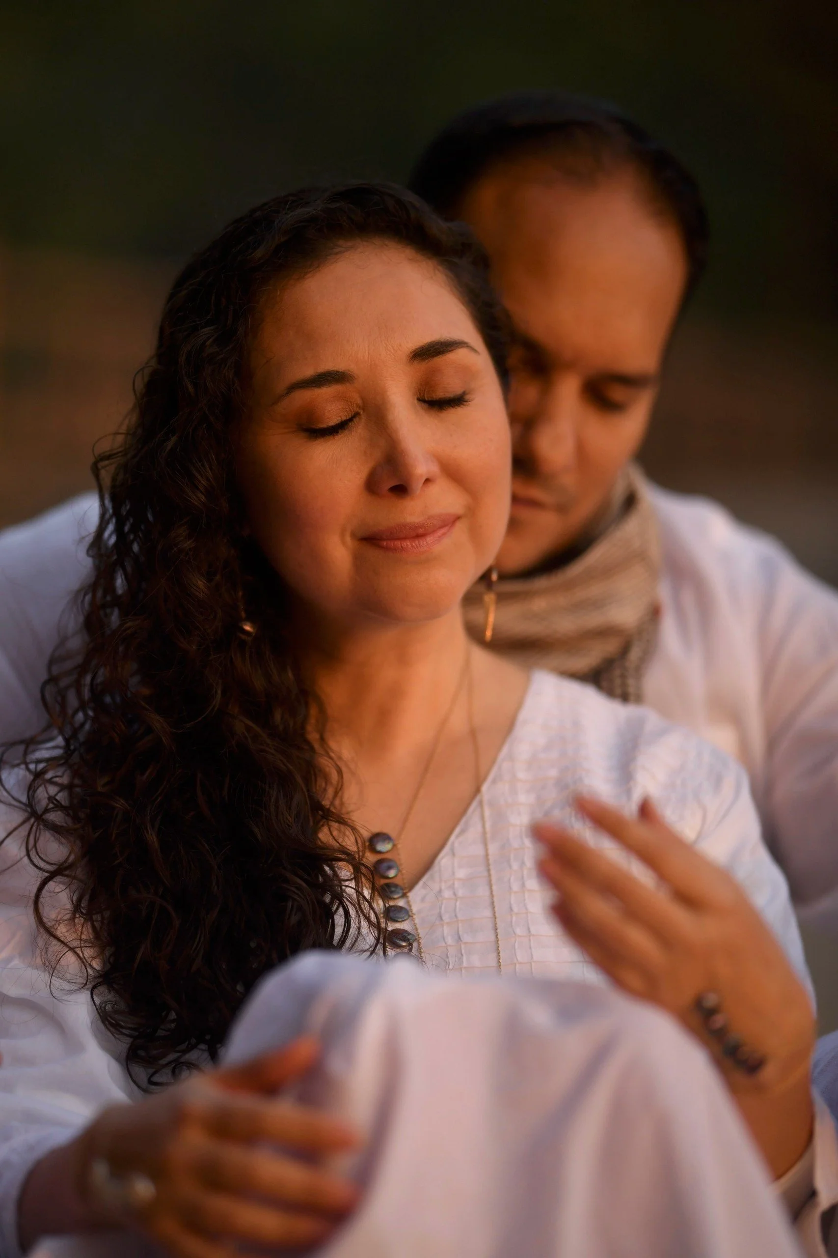 Woman with curly hair and eyes closed, wearing a ruffled top, posed indoors with a couch and plants in the background.