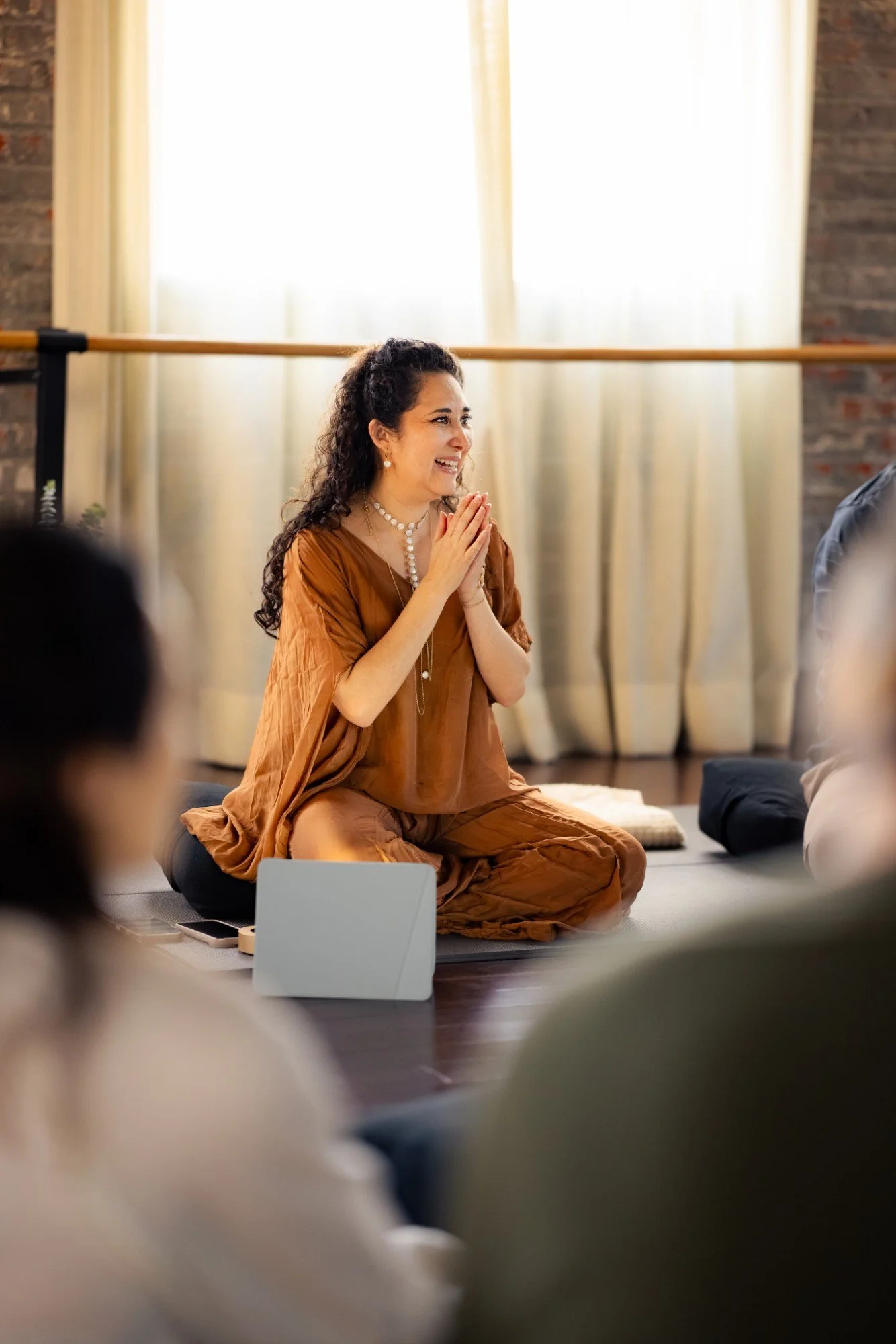 Woman sitting cross-legged on a cushion, smiling with hands clasped in front of a group, during a meditation or mindfulness session in a bright room with large windows and curtains.