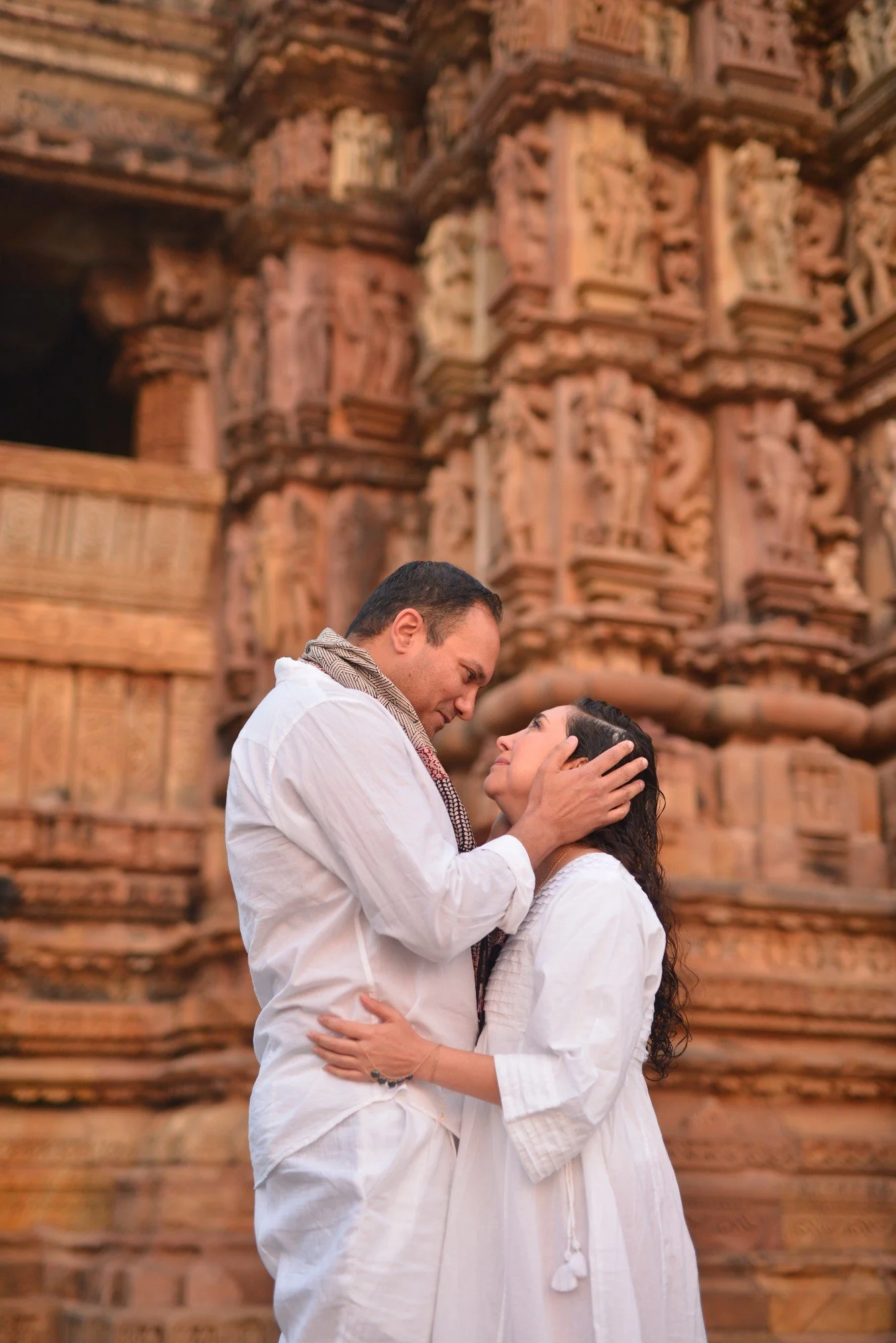 Woman with long dark hair standing near water, wearing a brown dress, eyes closed, hand on her chest.