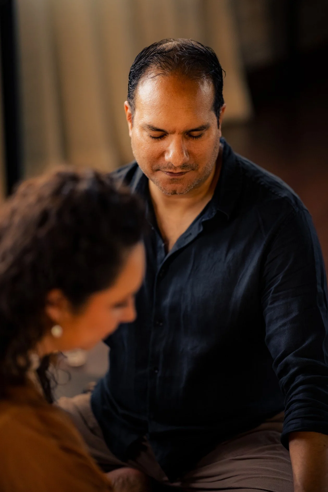 Woman with long dark hair standing near water, wearing a brown dress, eyes closed, hand on her chest.