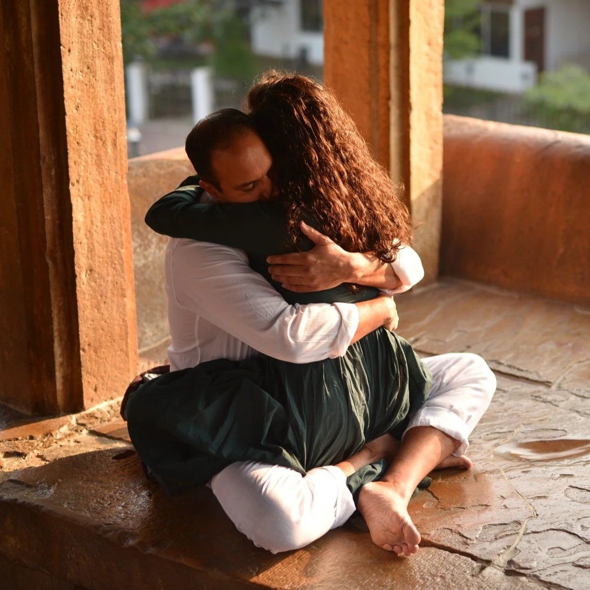 A woman with curly hair is sitting on a couch, writing in a notebook. She is wearing a beige dress and jewelry, and is surrounded by decorative pillows and indoor plants. A window is visible in the background.