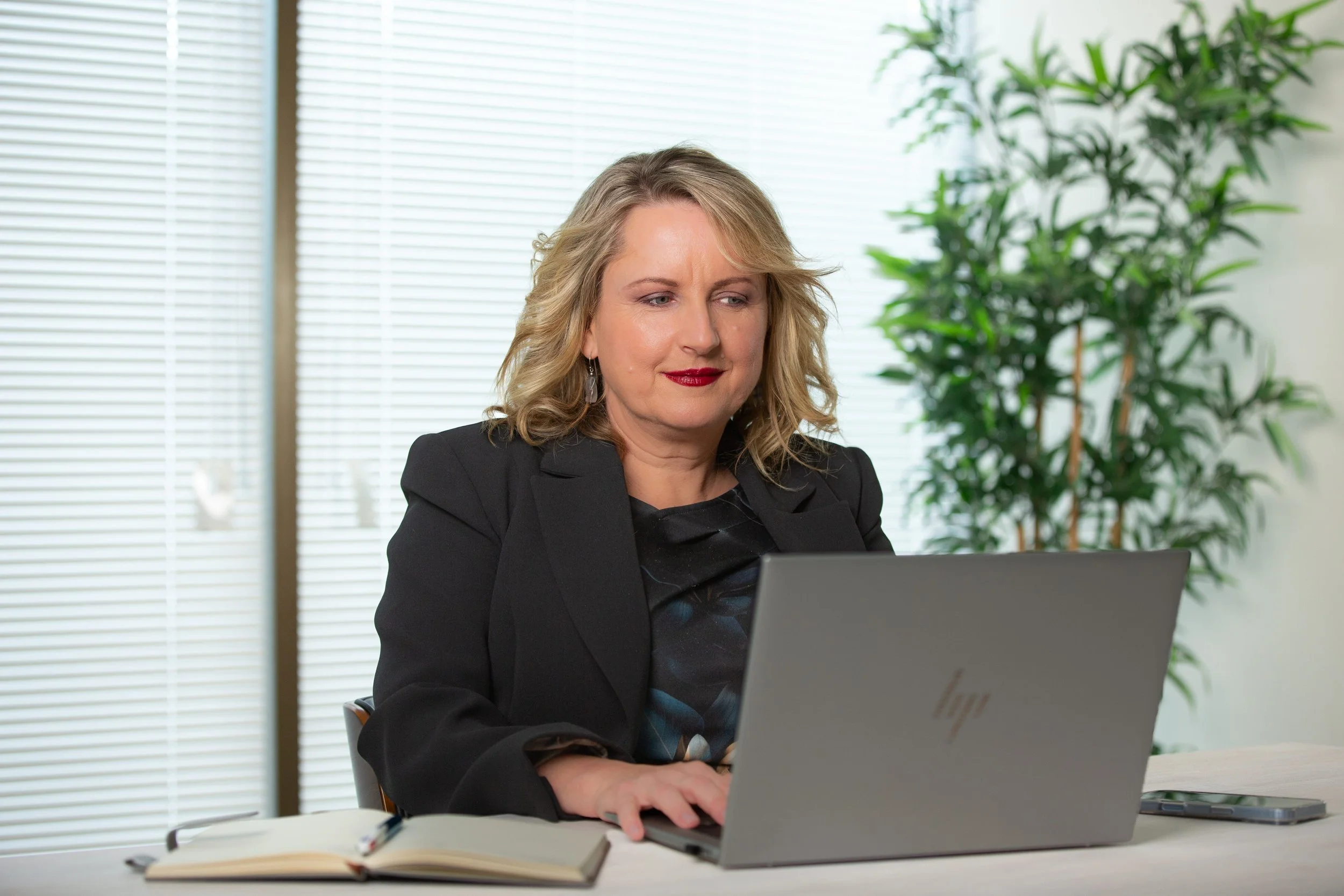 Ella Legge of The Story Was seated at a desk, working on a laptop in a modern office with large windows and green plants.