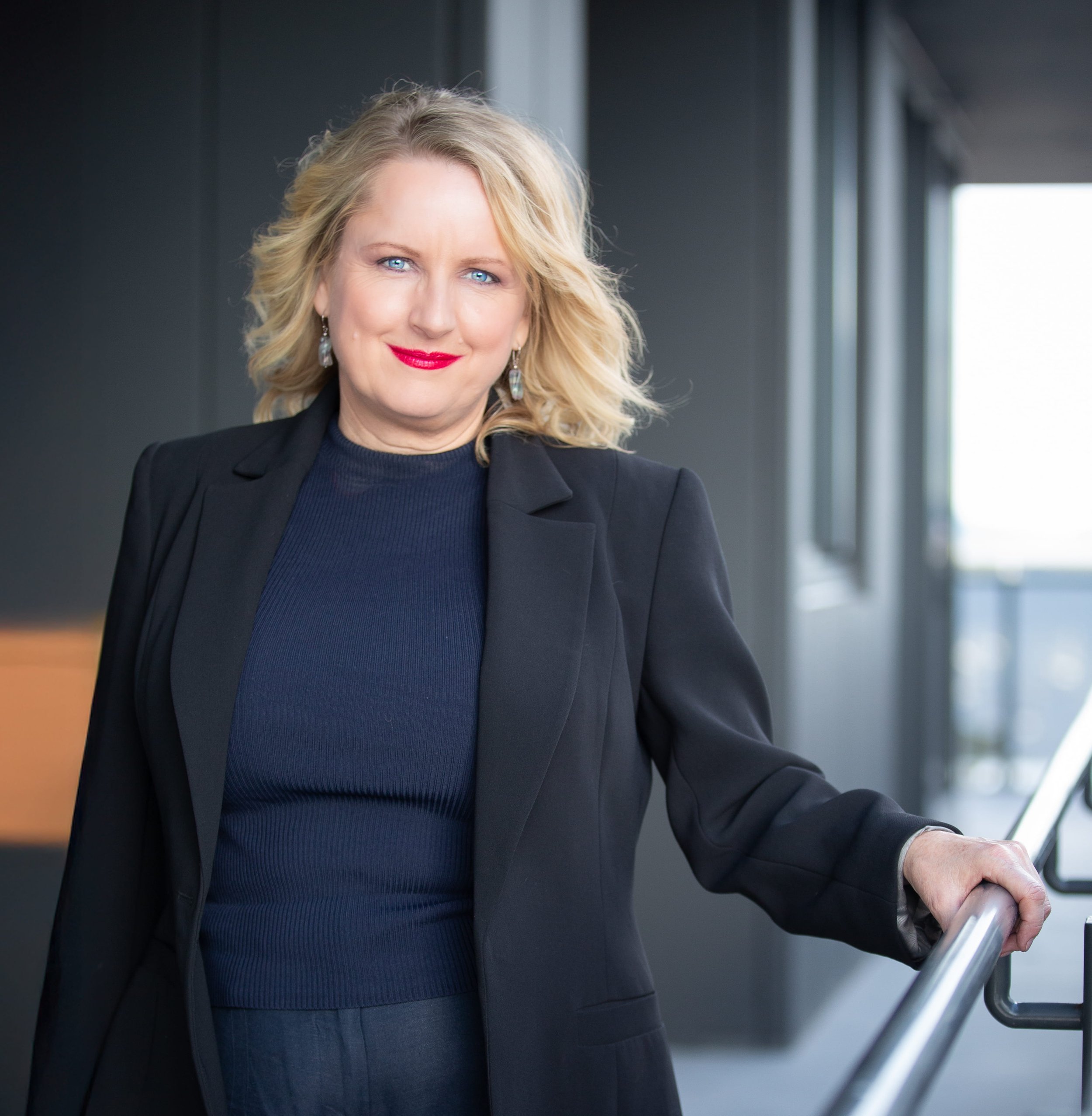 Ella Legge of The Story Was, wearing a navy blue top and black blazer, smiling confidently while standing in a modern indoor corridor with large windows.
