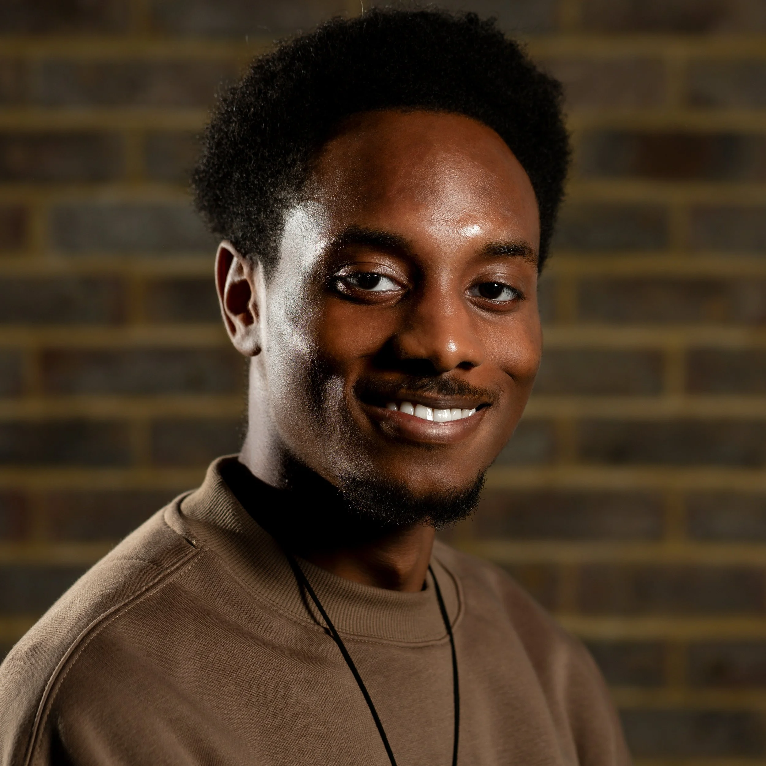 Close-up portrait of Kevon Anokwawe (OctoArtstuff), a young man with dark skin, short curly hair, smiling, wearing a brown shirt and a black necklace, standing in front of a brick wall background.