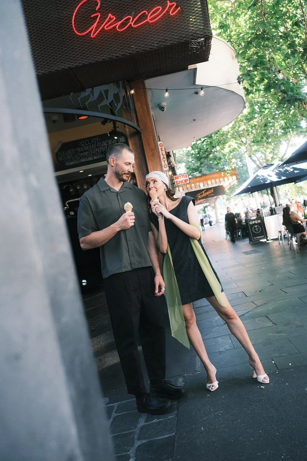 A groom and bride each holding an ice cream cone. The groom is smiling at the bride while she looks at him playfully. She is dressed in a black dress with green sheer panels and high-heeled shoes, and the groom is wearing a dark shirt and pants.