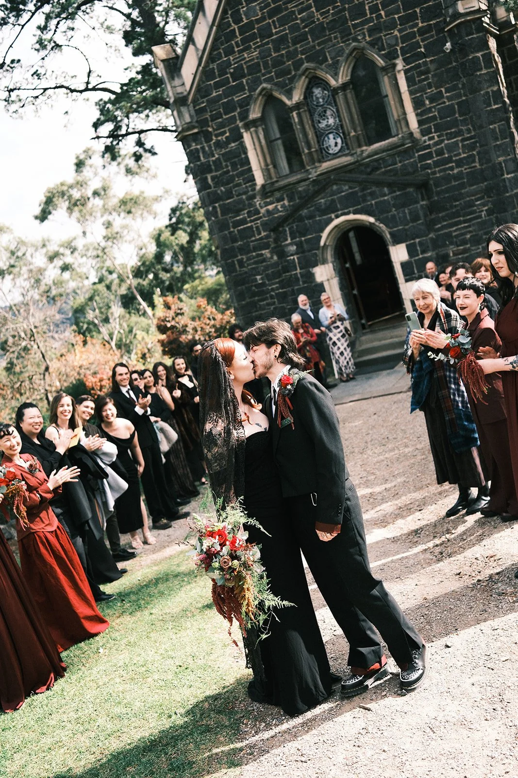 A bride and groom share a kiss after theoir ceremony, outside the chapel at Montsalvat in the Yarra Valley. Their guests are stood behind them clapping. 