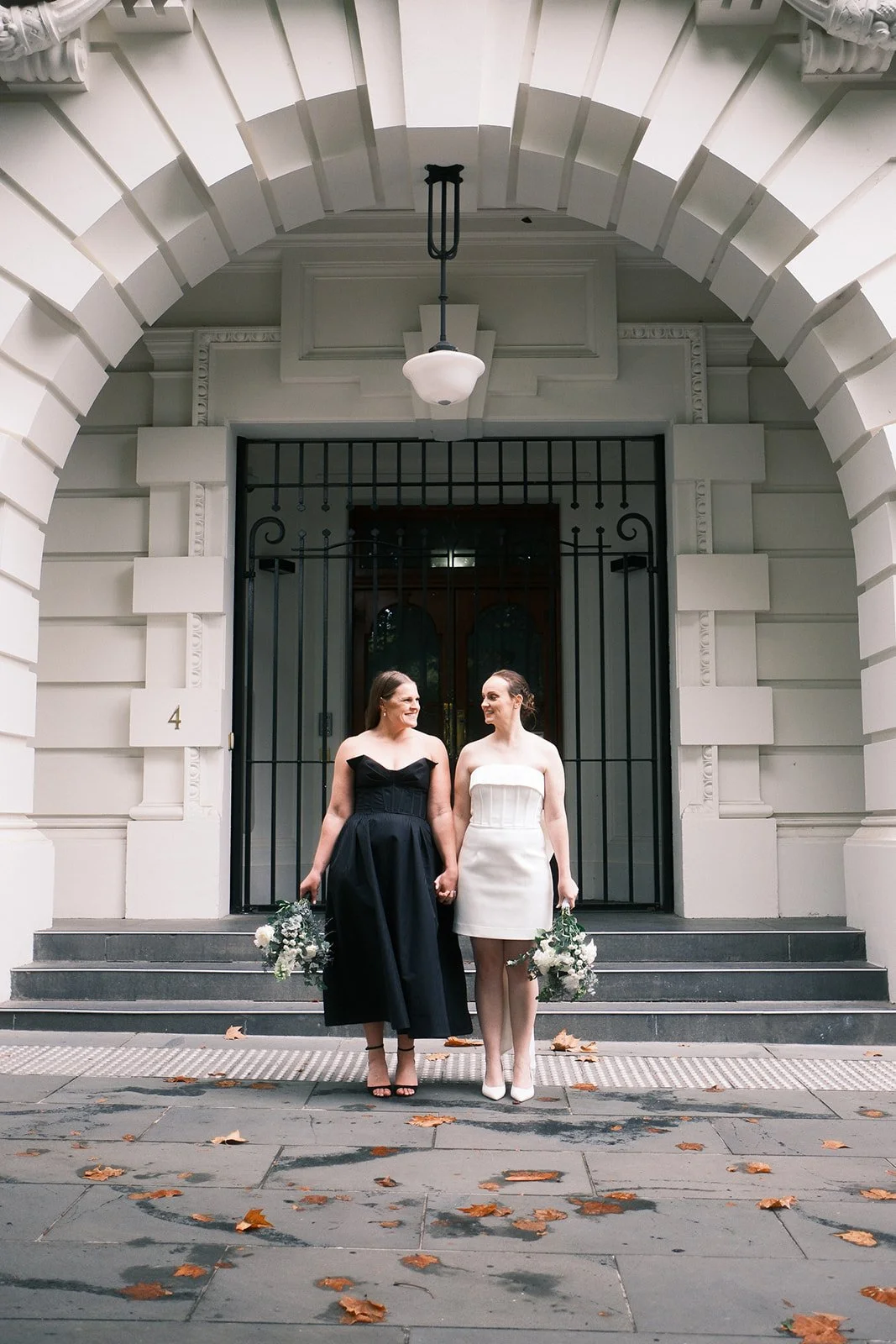 Two women in wedding dresses holding hands, one in a black dress and the other in a white dress, standing on steps outside a building with an arched entrance.
