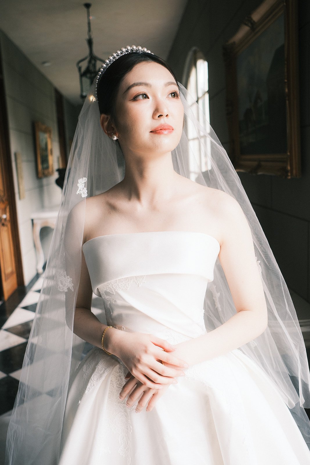 A bride in a strapless white wedding dress with a pearl headband and veil, standing indoors near a window, with artwork on the wall behind her at Campbell Point House.