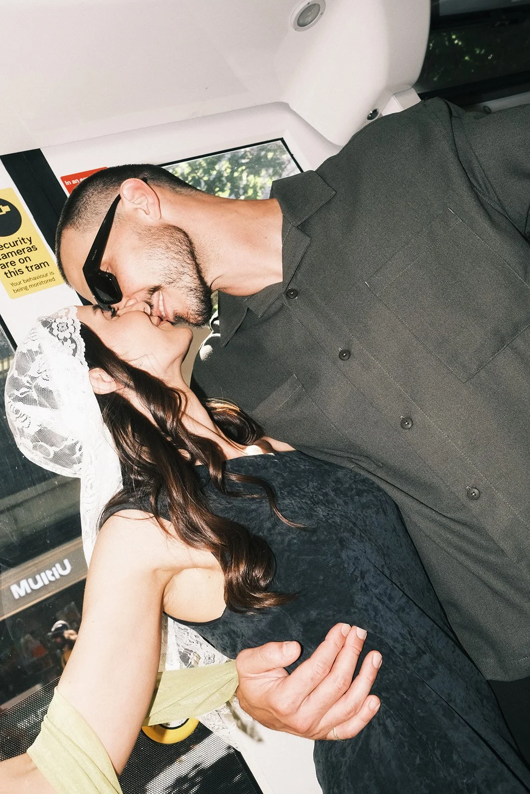A groom and bride share a kiss on a tram after their elopement ceremony at the Victorian Marriage Registry. The woman wears a lace head covering and a sleeveless top, while the man wears sunglasses and a button-up shirt.