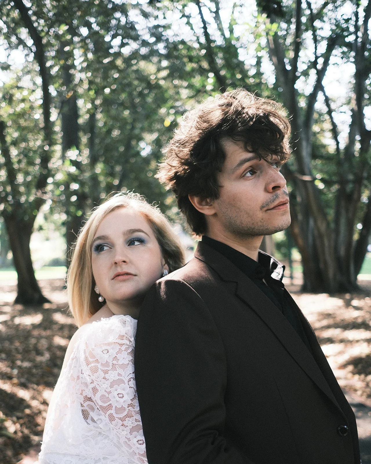A groom and a bride standing back-to-back in the Treasury Gardens in Melbourne, with trees and sunlight in the background. The bride is resting her head on her groom's shoulder, and they both have serious expressions.