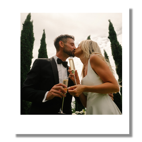 A bride and groom kissing outdoors during their wedding, holding champagne glasses.