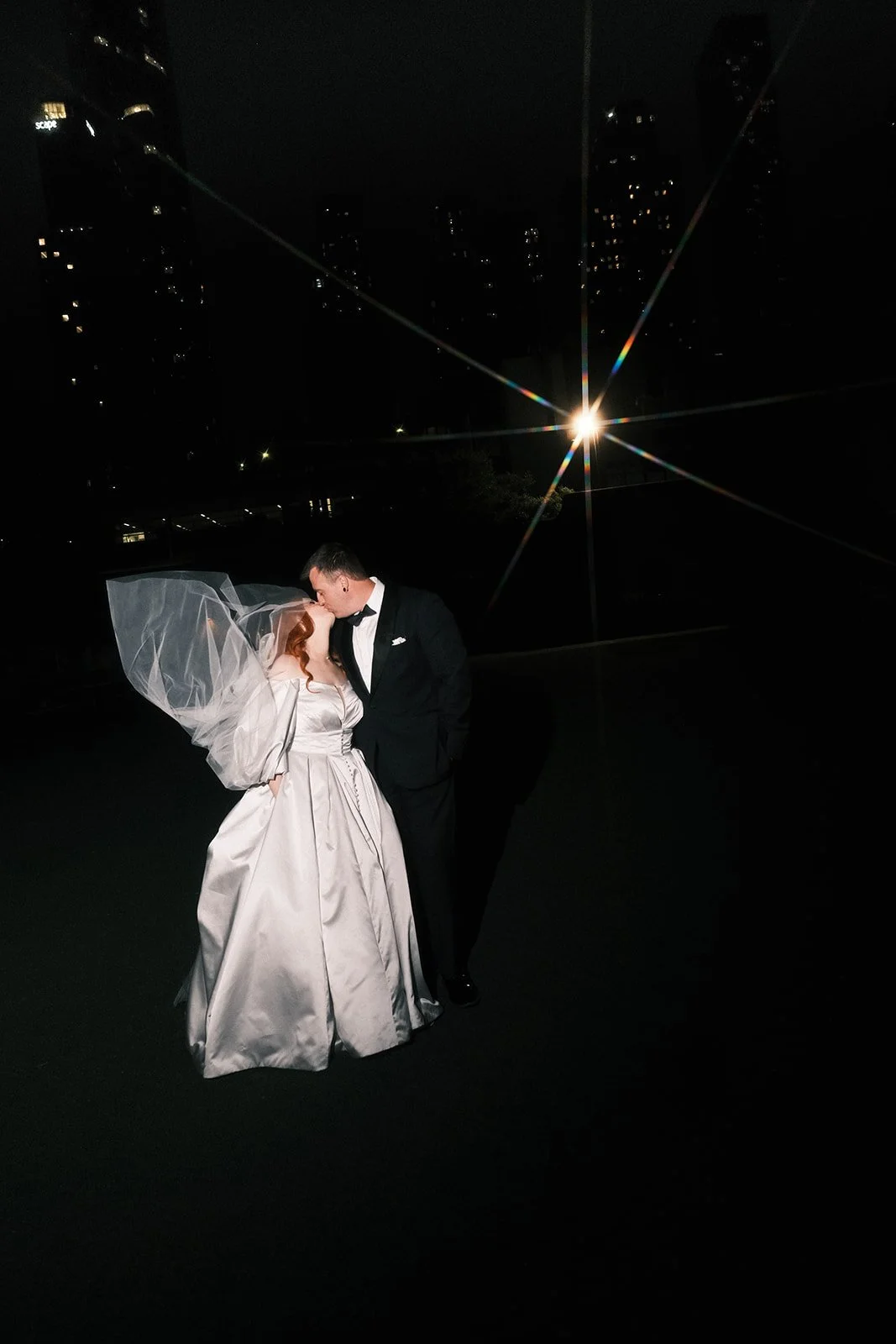 A bride and groom share a kiss at night, with a Melbourne city skyline and bright light in the background.