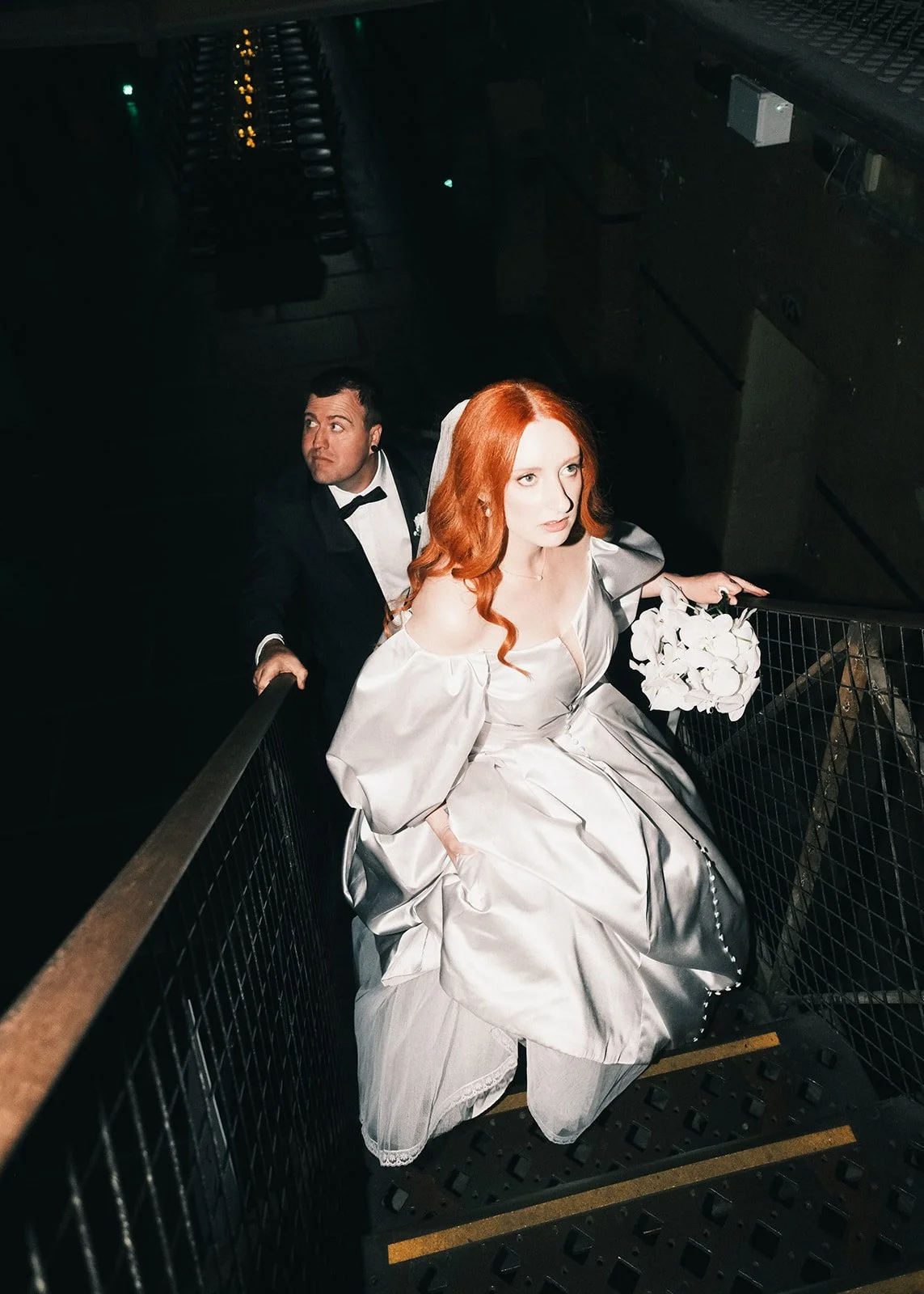 A woman with red hair in a wedding dress holding a bouquet, climbing a staircase behind a man in a tuxedo, with a dark background.