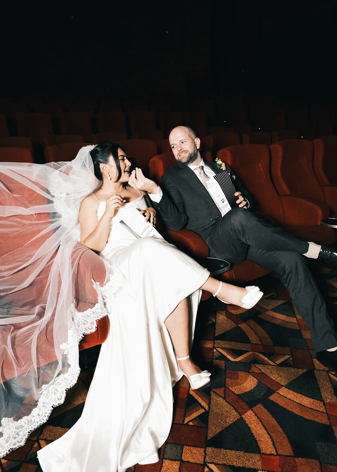 A bride and groom feeding each other popcorn after their wedding ceremony at Palace Cinemas Westgarth. They are sitting in the cinema theatre chairs, smiling.