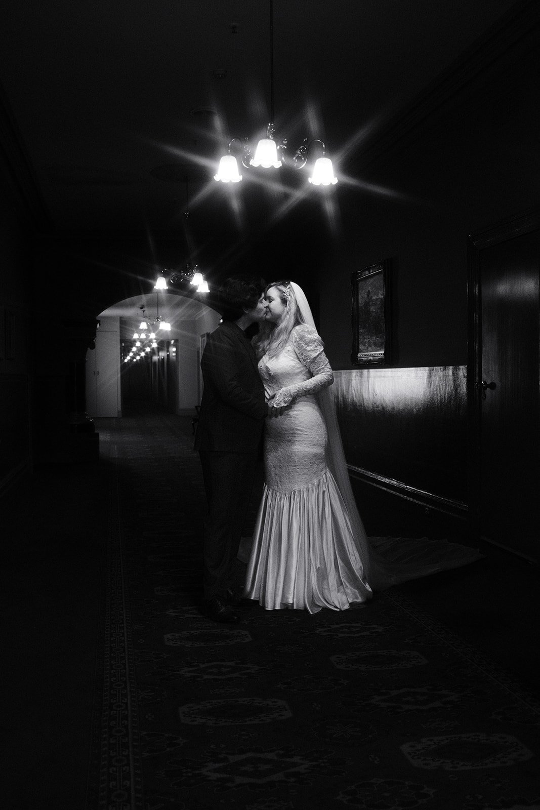 A black and white photo of a bride and groom sharing a kiss in the dimly lit hallway of The Hotel Windsor, with hanging chandeliers.