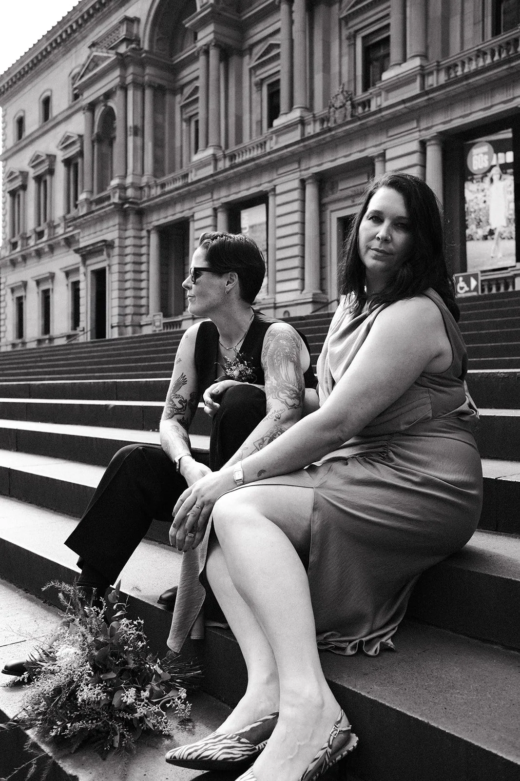 Two brides sitting on the steps of the Old Treasury Building after their wedding ceremony at the Victorian Marriage Registry.
