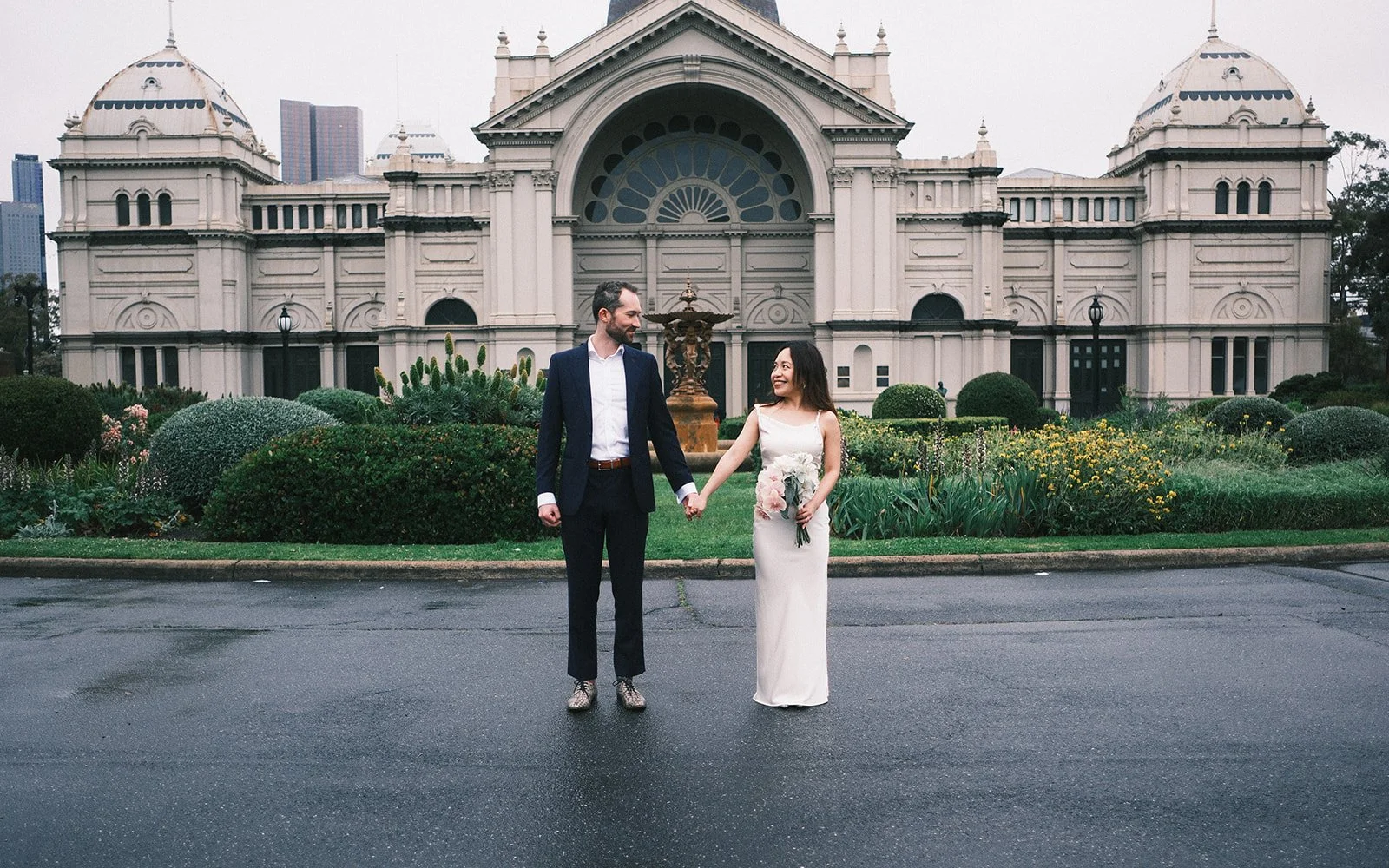 A couple holding hands on their engagement session in Melbourne. They are standing in front of the Royal Exhibition Buildings in Melbourne, looking at each other.