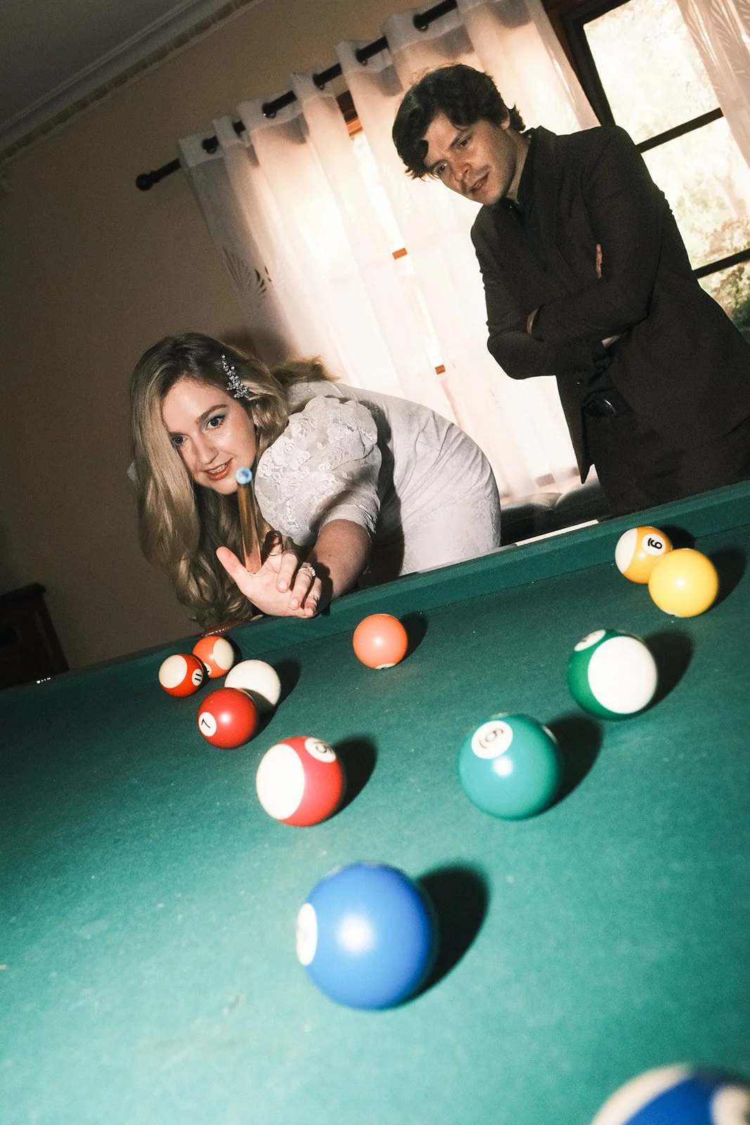 A bride playing pool before her wedding, with the groom standing nearby, observing. The pool balls are scattered on the green felt surface of the table.