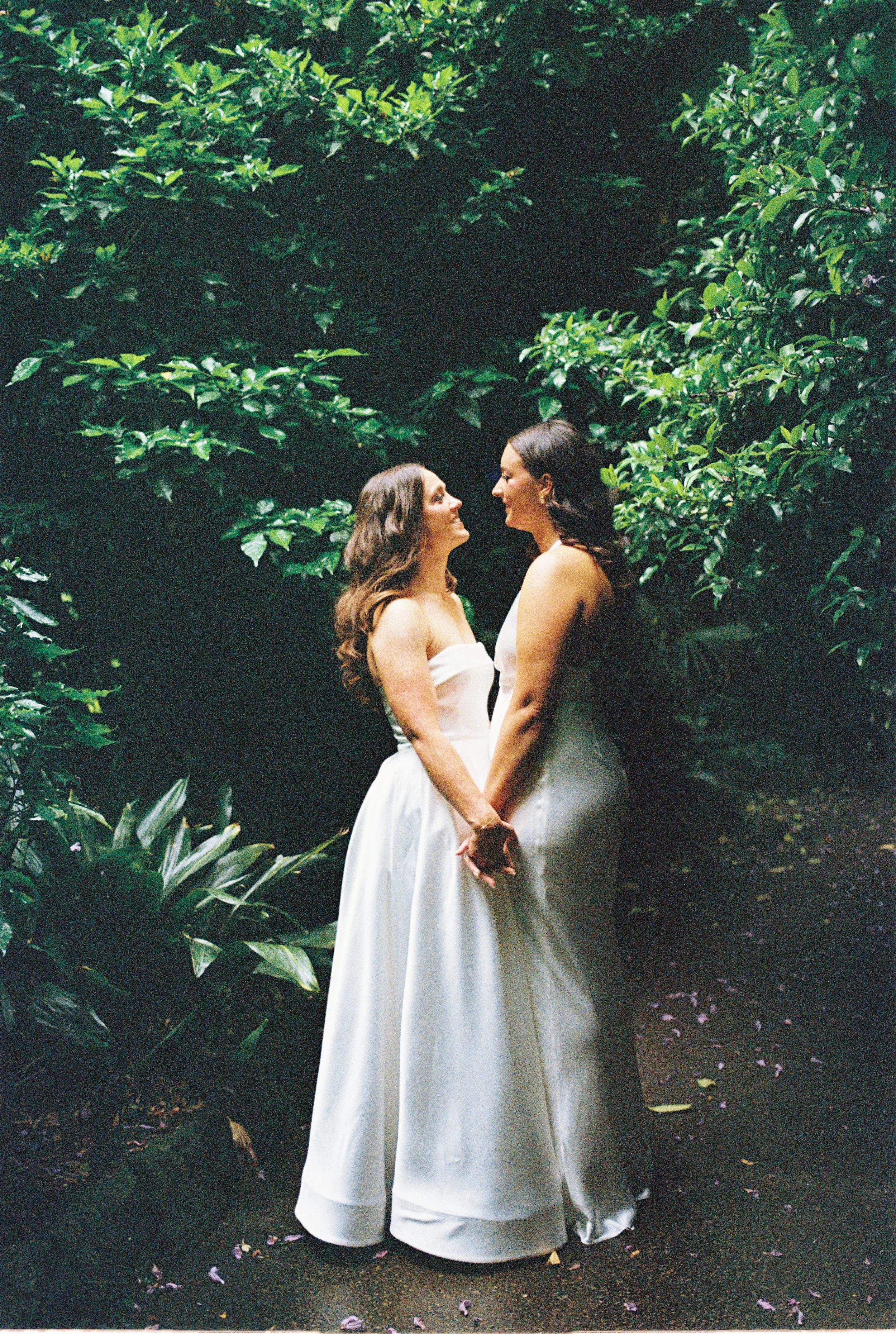 Two brides in wedding dresses standing face to face, holding hands, surrounded by lush green foliage in a garden setting.