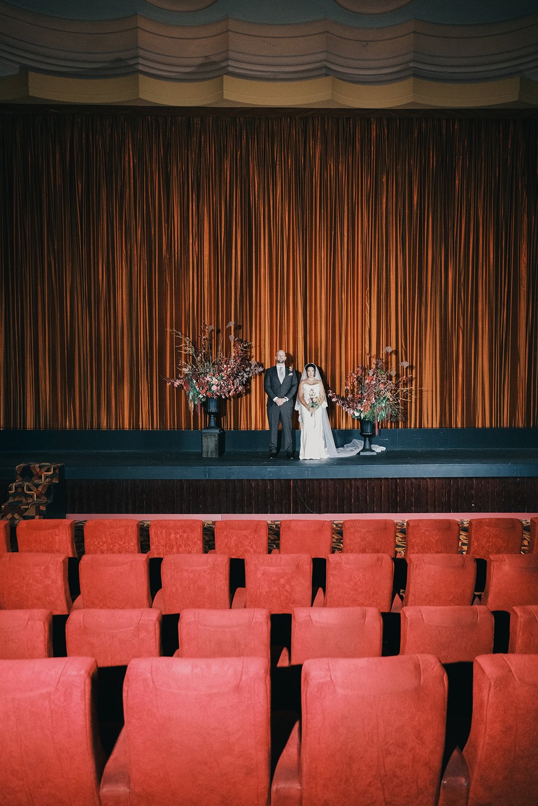 A bride and groom stand on stage at Westgarth Palace cinema in Northcote during a wedding ceremony with large flower arrangements on either side, in front of dark orange curtains, in an cinema with red velvet seats.