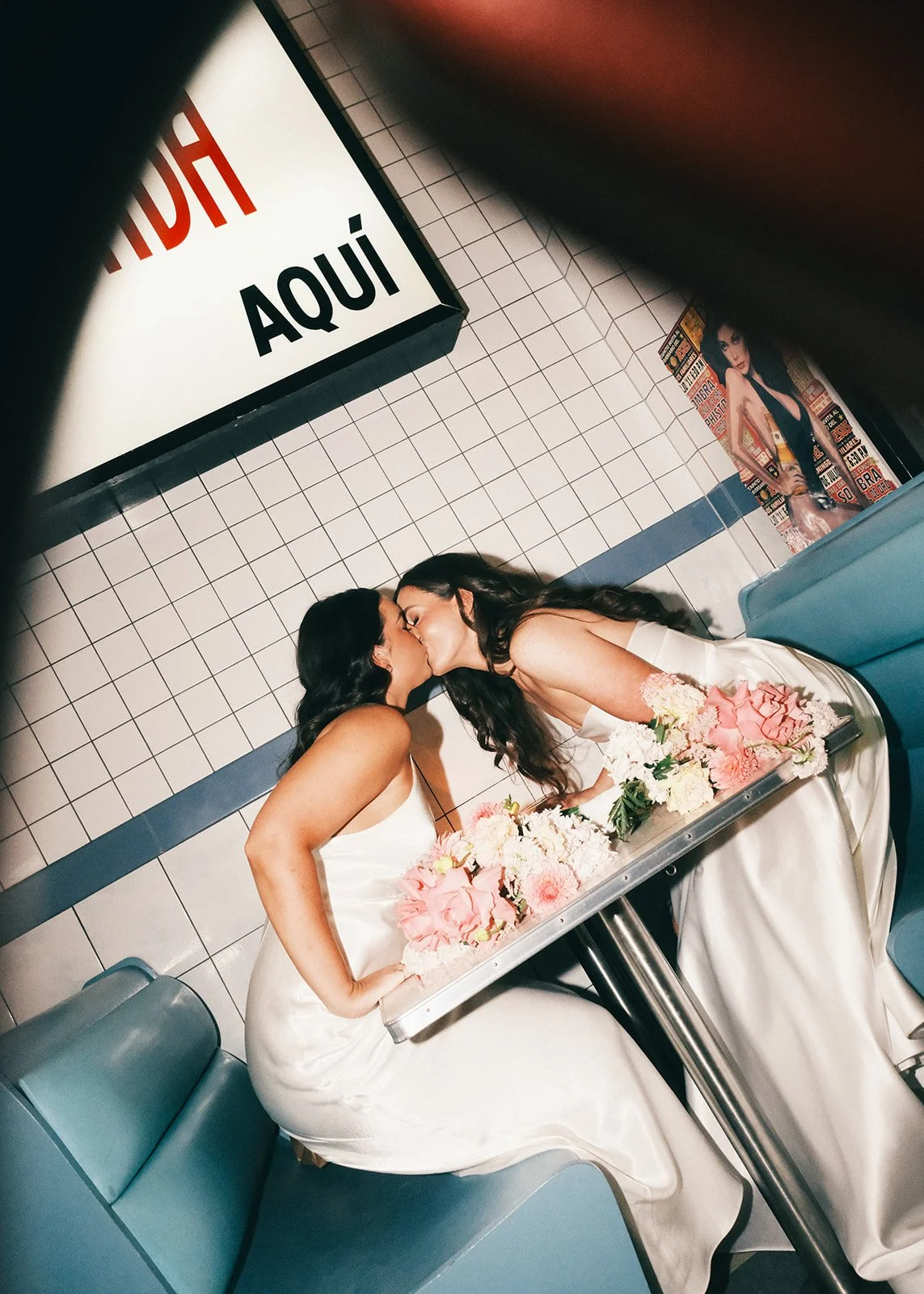 Two women in wedding dresses sharing a kiss in a diner booth with floral decorations on the table.