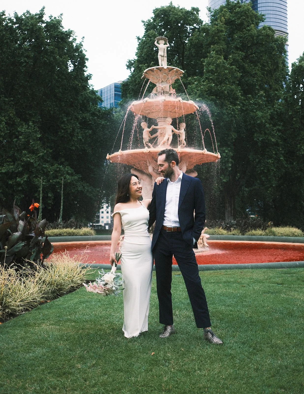 A man and woman on their engagement shoot in Carlton Gardens. They are standing in front of a water fountain, looking at each other. He is wearing a blue suit, while she is wearing a white wedding dress.