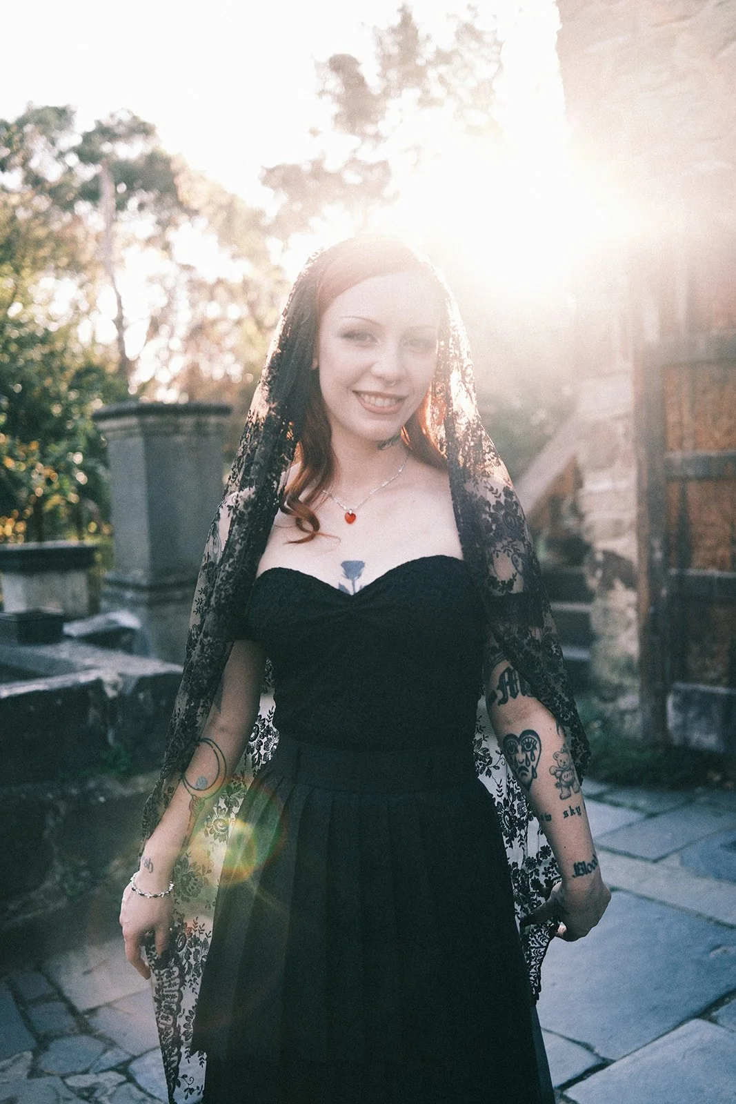 A bride at her wedding at Montsalvat in the Yarra Valley. She is wearing a non-traditional black dress and black veil, and the sun is setting behind her.