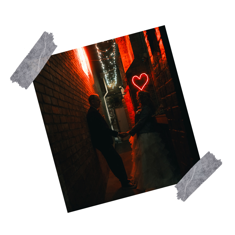 A bride and groom, holding hands in a dimly lit alley at The Hall at Welcome To Brunswick with red neon and string lights, and a heart-shaped neon sign in the background.