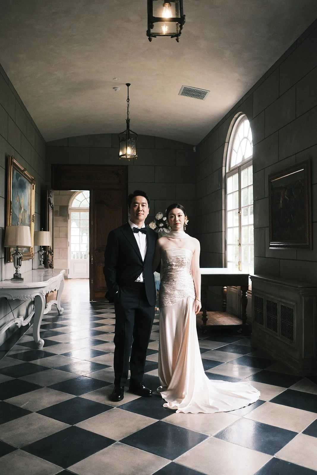 A couple dressed in formal wedding attire standing inside Campbell Point House. The large windows light them up.