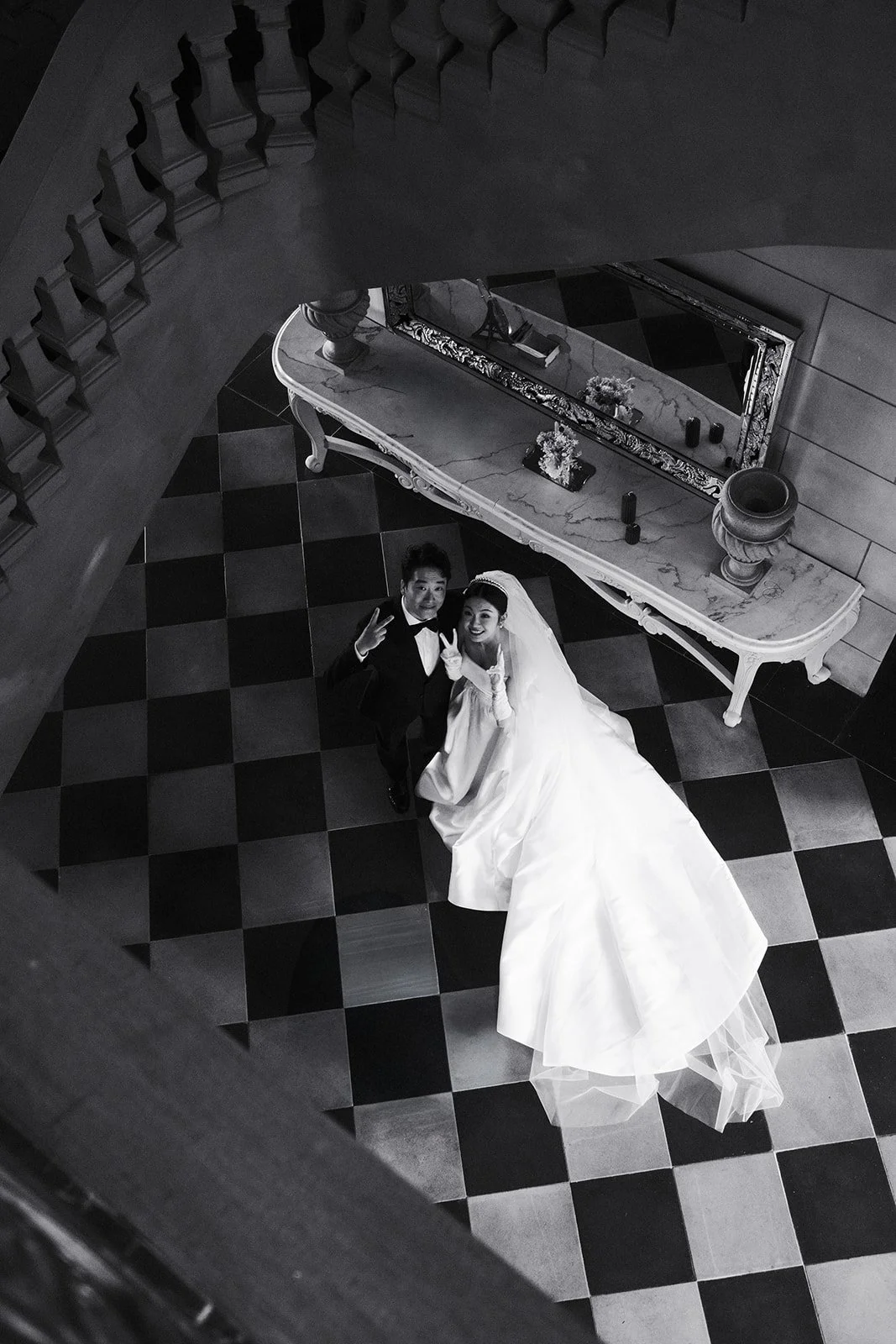 Black and white photo of a bride and groom looking up at the camera inside the hallway of Campbell Point House. The bride is wearing a wedding dress with a veil, and the groom is in a tuxedo.