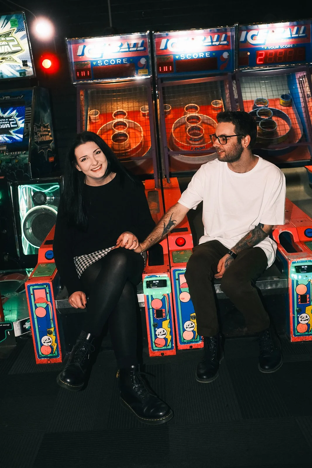 A couple at an arcade, posing for engagement photos. They are sitting on the arcade machine, holding hands.