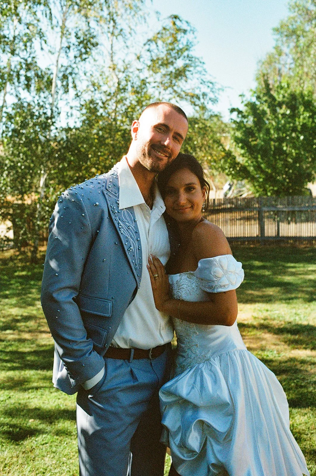 A bride and groom in wedding attire standing close together at a private property wedding on a sunny day, smiling, with trees and flowers in the background.