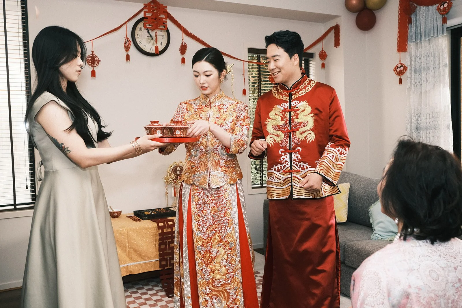 A traditional Chinese wedding tea ceremony with the couple in embroidered attire, offering tea to a guest in a beige dress, indoors with red and gold decorations.