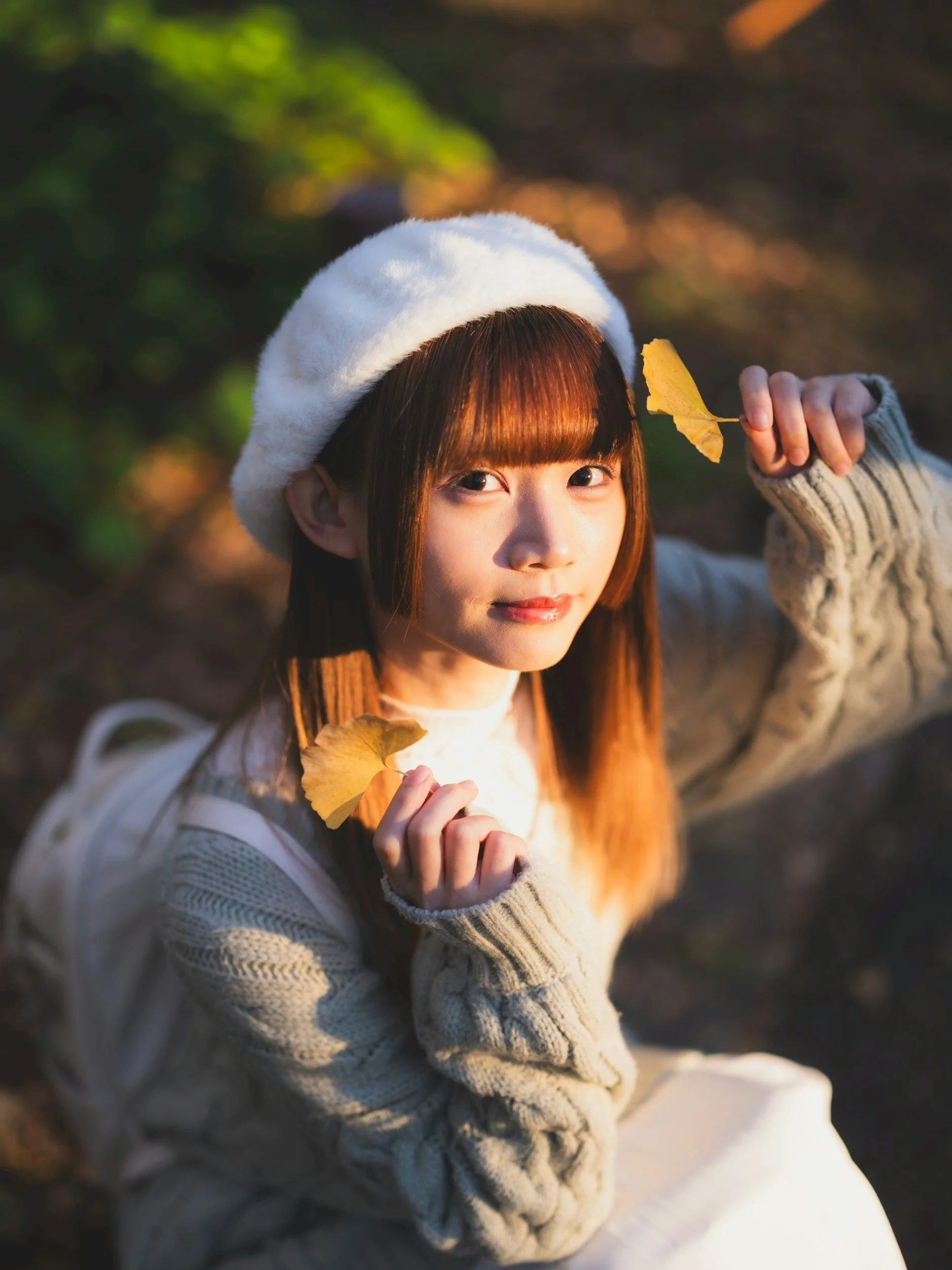 A young woman with long brown hair and bangs, wearing a white beret and a beige knit sweater, holding yellow autumn leaves near her face, outdoors in a forest.